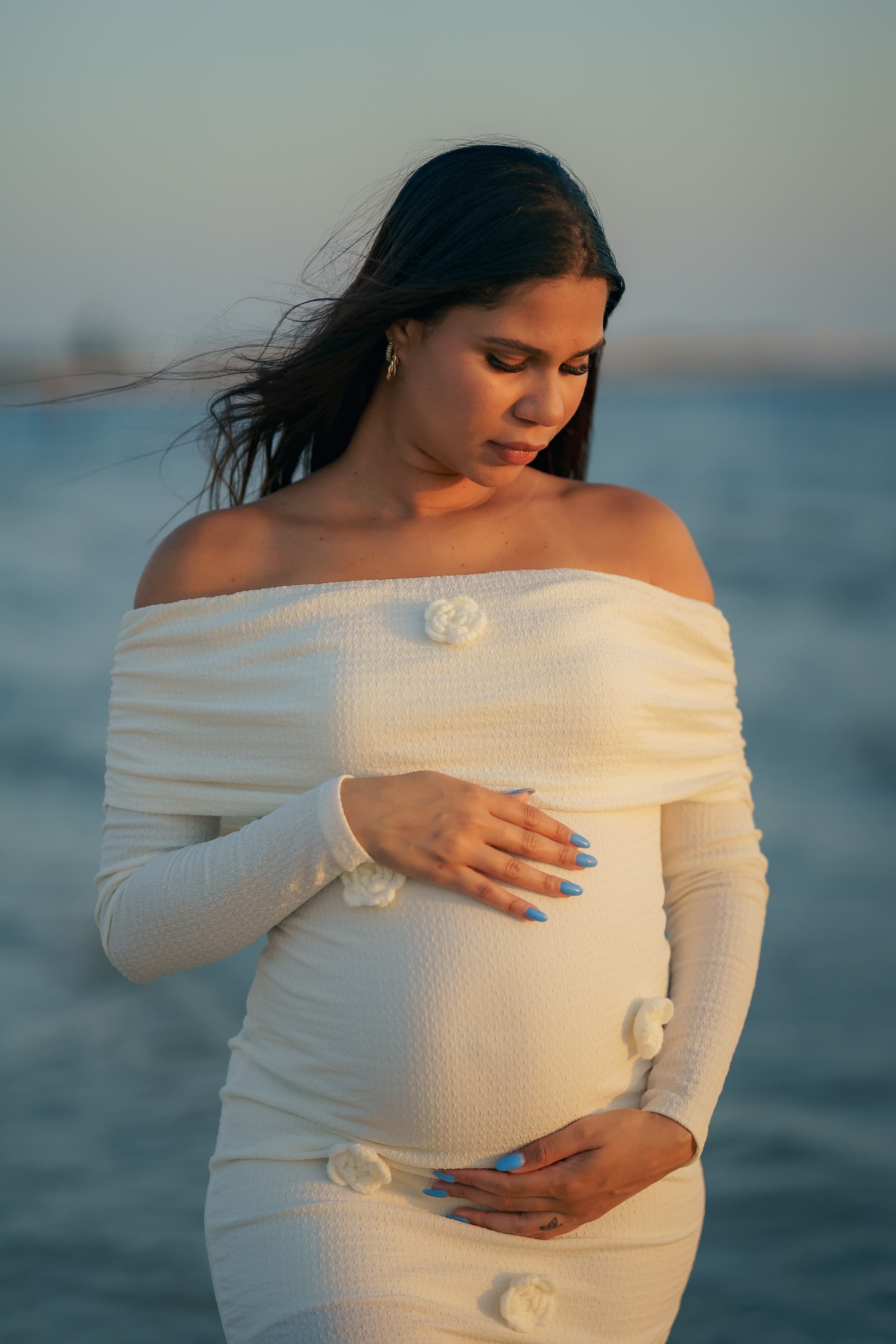 A pregnant woman in a white off-shoulder dress stands by the ocean, gently touching her belly and looking down, with her hair blowing in the wind during sunset. Cinematic Editorial Pictures in Tampa Florida.