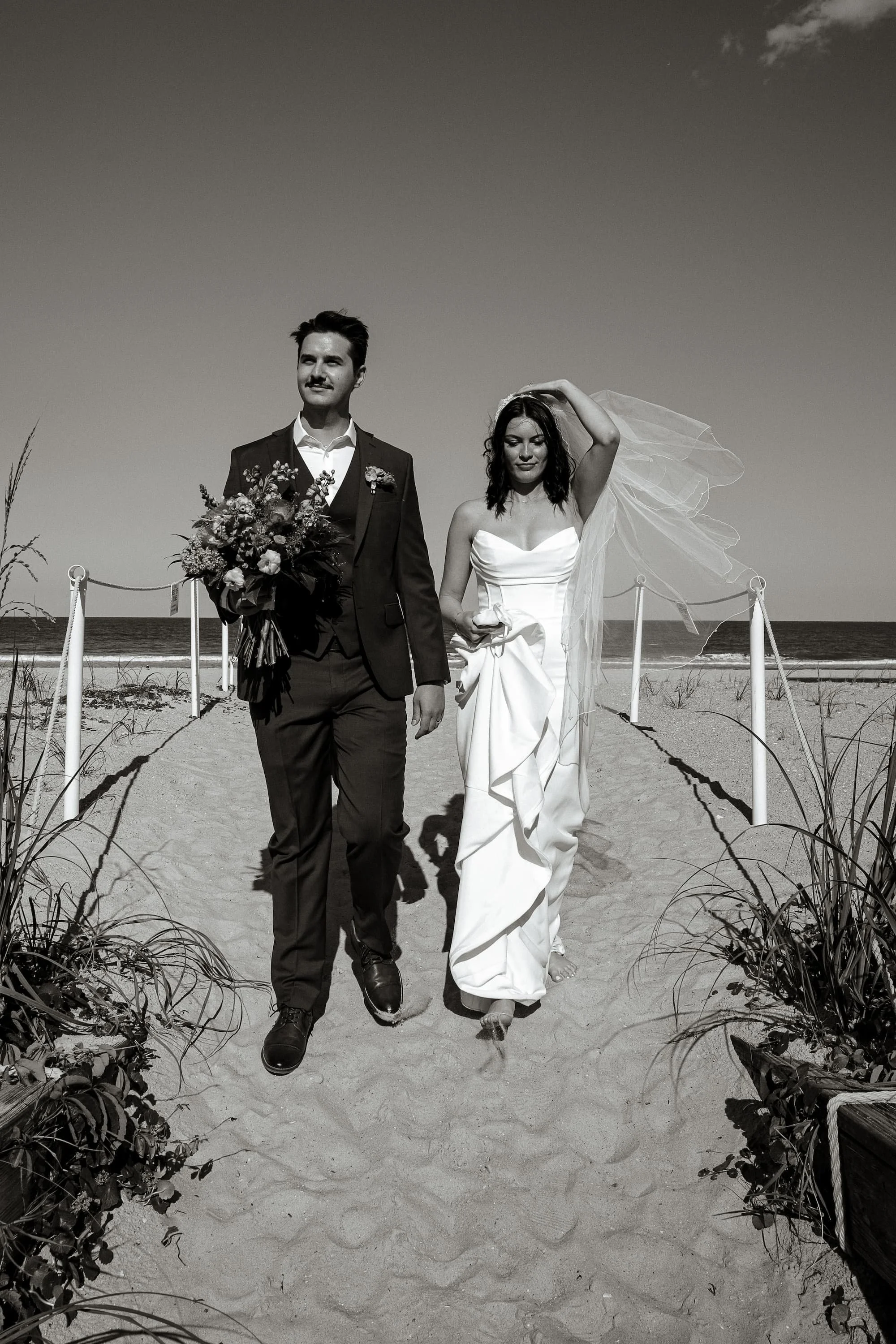 Black and white photo of a bride and groom walking on a sandy beach aisle, with the ocean in the background. The groom is in a suit holding a bouquet, and the bride is in a wedding dress with a veil, holding the dress with one hand.Cinematic wedding