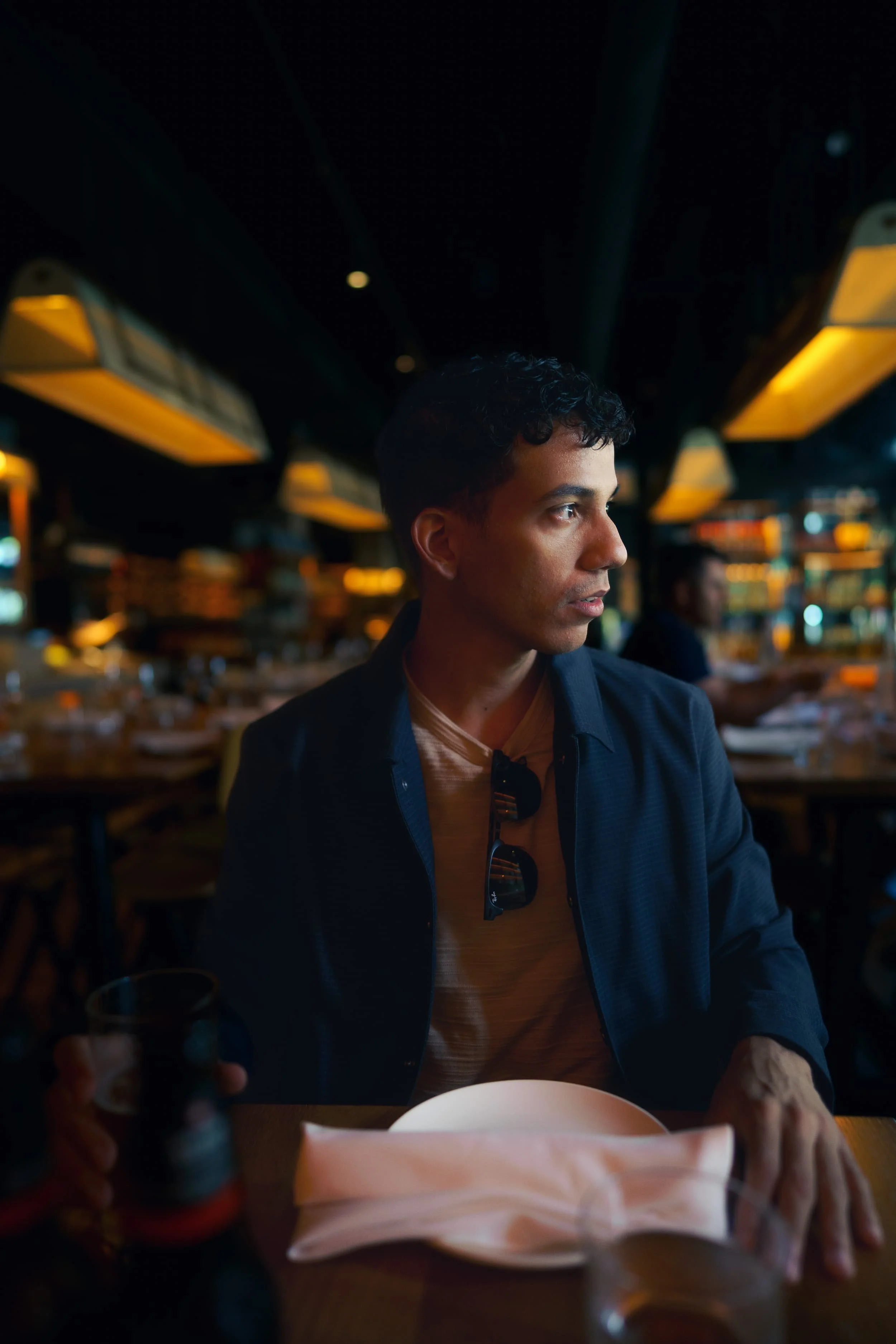 A young man with short curly hair sits at a table in a dimly lit restaurant, looking to his right with sunglasses hanging from his shirt collar. He is wearing a navy jacket and a beige shirt. Cinematic Editorial Pictures in Tampa Florida.