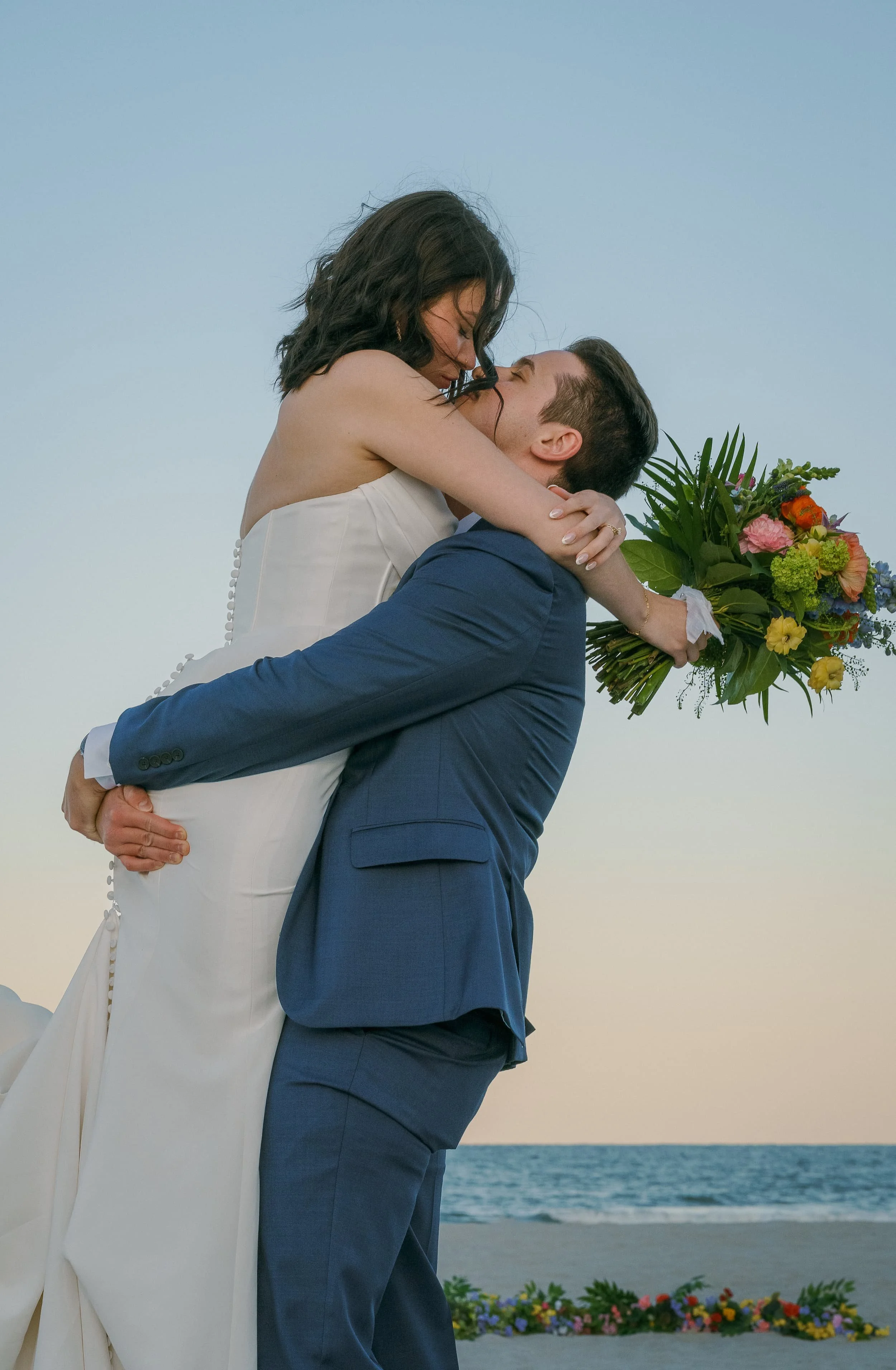 A couple in wedding attire embracing and about to kiss on the beach during sunset. The bride is holding a colorful bouquet of flowers. Cinematic Editorial Pictures in Tampa Florida.