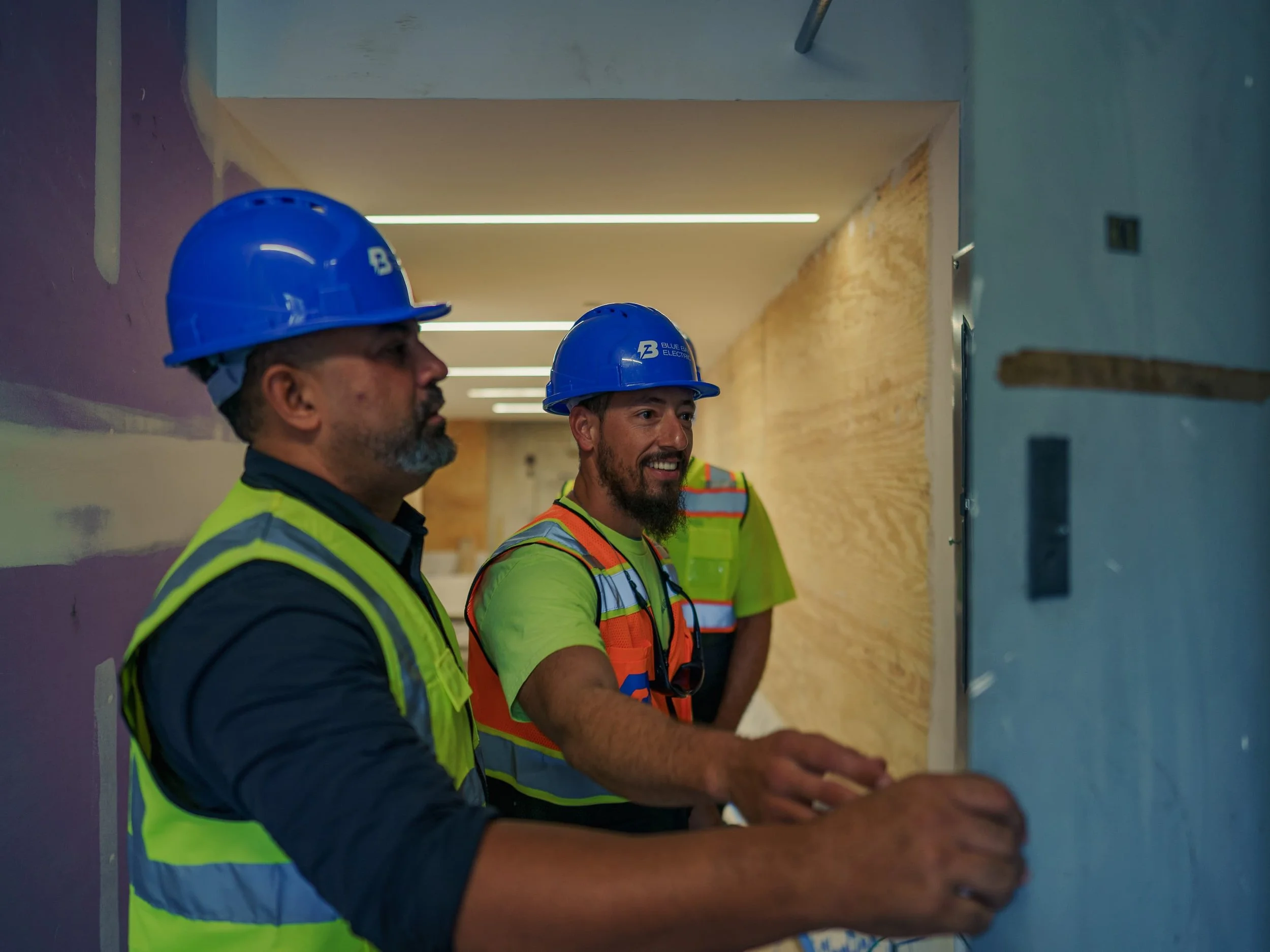Two construction workers wearing blue hard hats and reflective safety vests working on an electrical panel in a building under construction. Cinematic Editorial Pictures in Tampa Florida. Small brand business on tampa florida