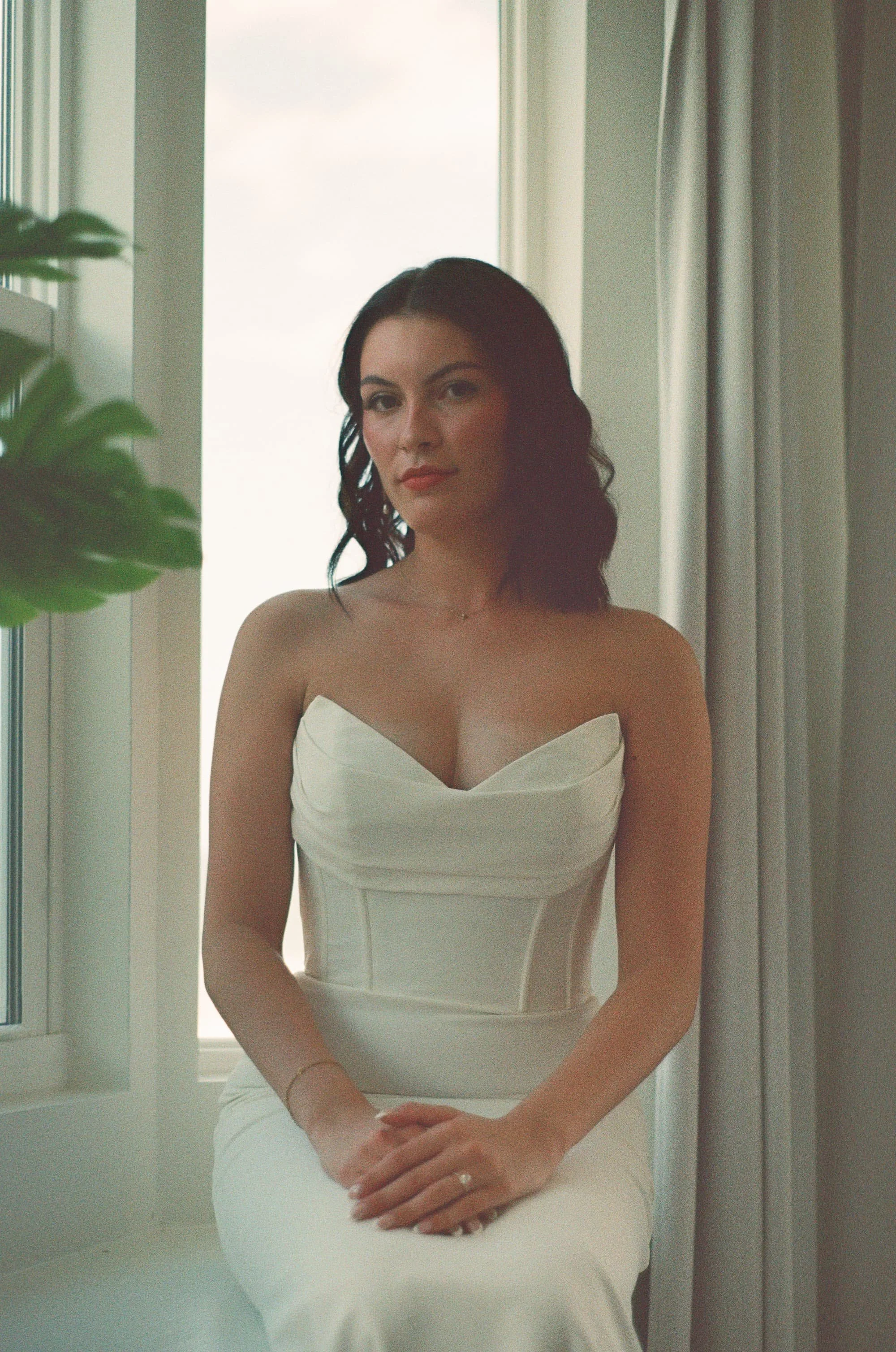 A woman with dark wavy hair and light skin, dressed in a strapless white gown, sitting by a window in a softly lit room. Cinematic Editorial Pictures in Tampa Florida.  Wedding Bride, White dress. Wedding getting ready.