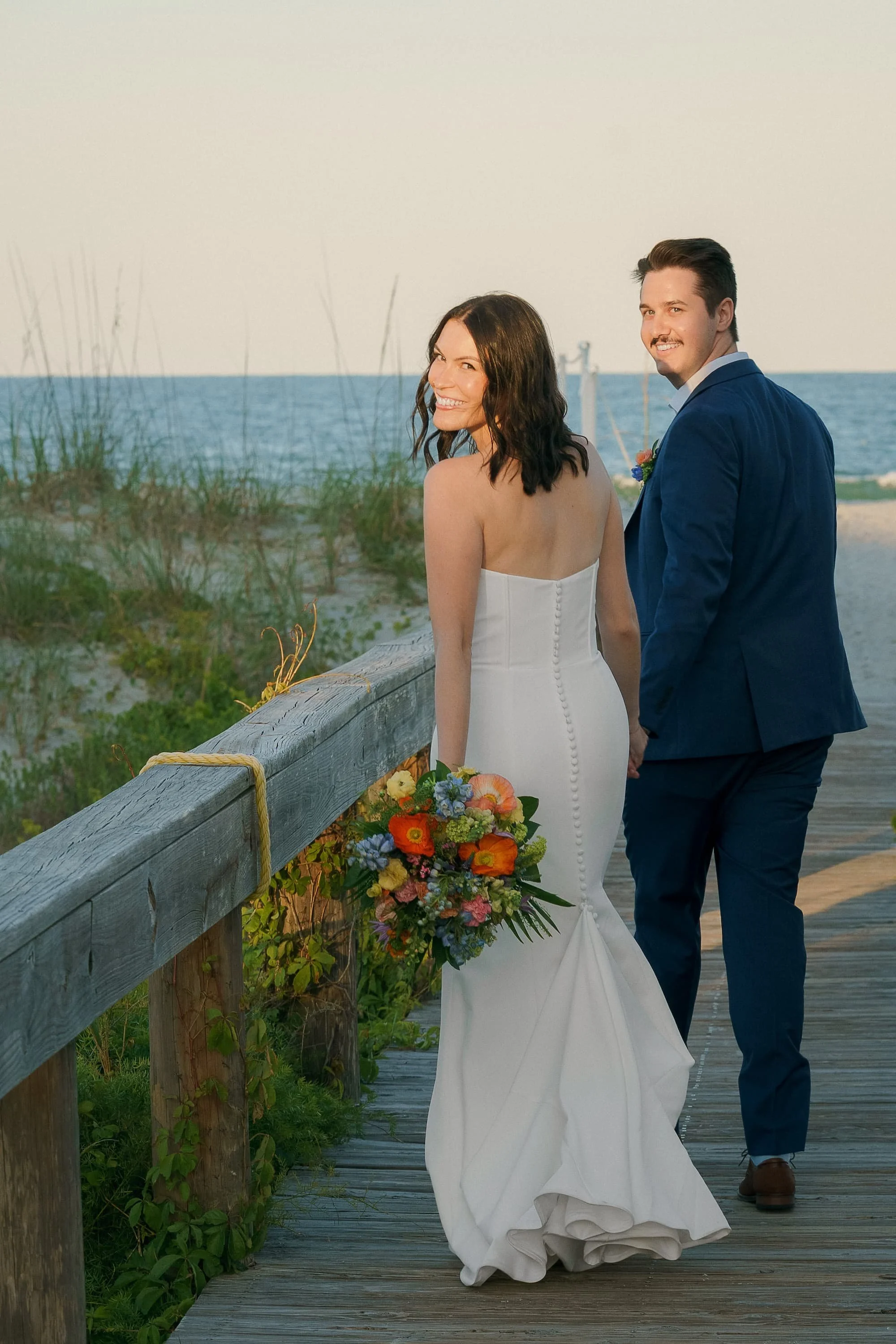A bride and groom holding hands on a beach boardwalk, smiling and looking back at the camera, with ocean and sky in the background. Cinematic Editorial Pictures in Tampa Florida.