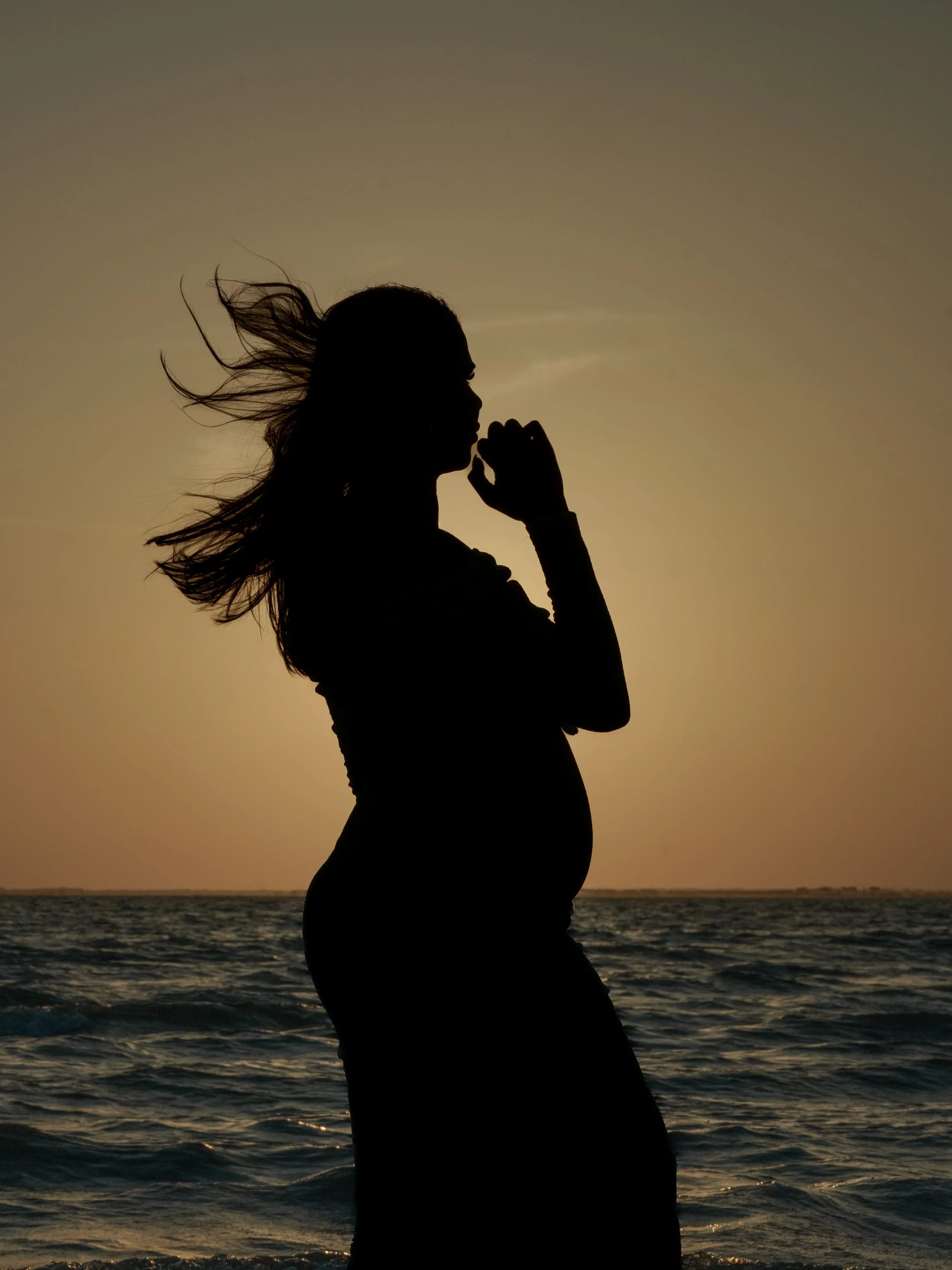 Silhouette of a pregnant woman standing by the ocean at sunset, with wind-blown hair and her hand near her face. Cinematic Editorial Pictures in Tampa Florida.