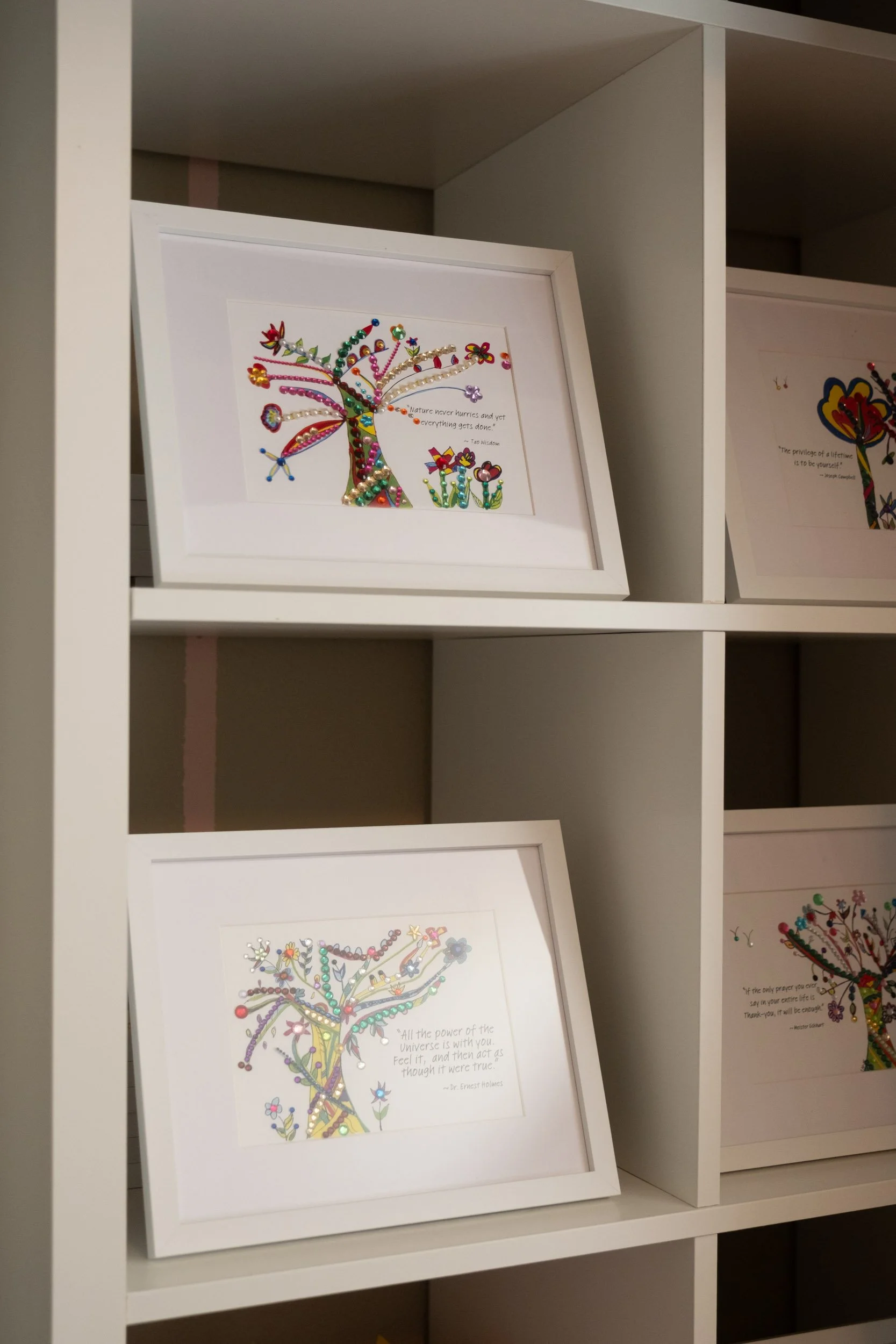 Framed artwork of trees with colorful beads and decorations placed on white shelves.