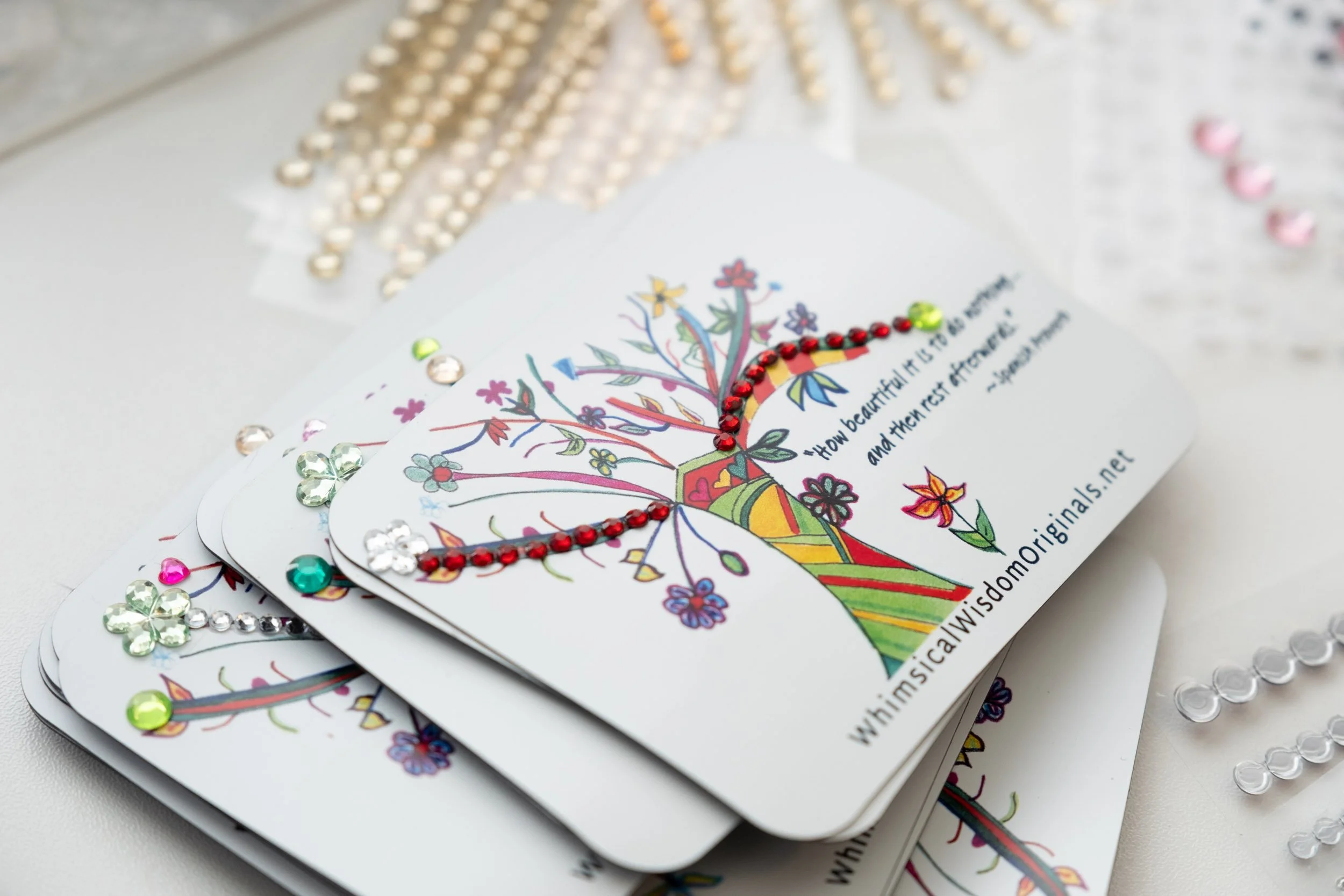 Stack of illustrated cards decorated with colorful rhinestones, featuring a drawing of a tree with flowers and a quote about beauty and rest. Additional decorative beads and sequins are visible in the background.