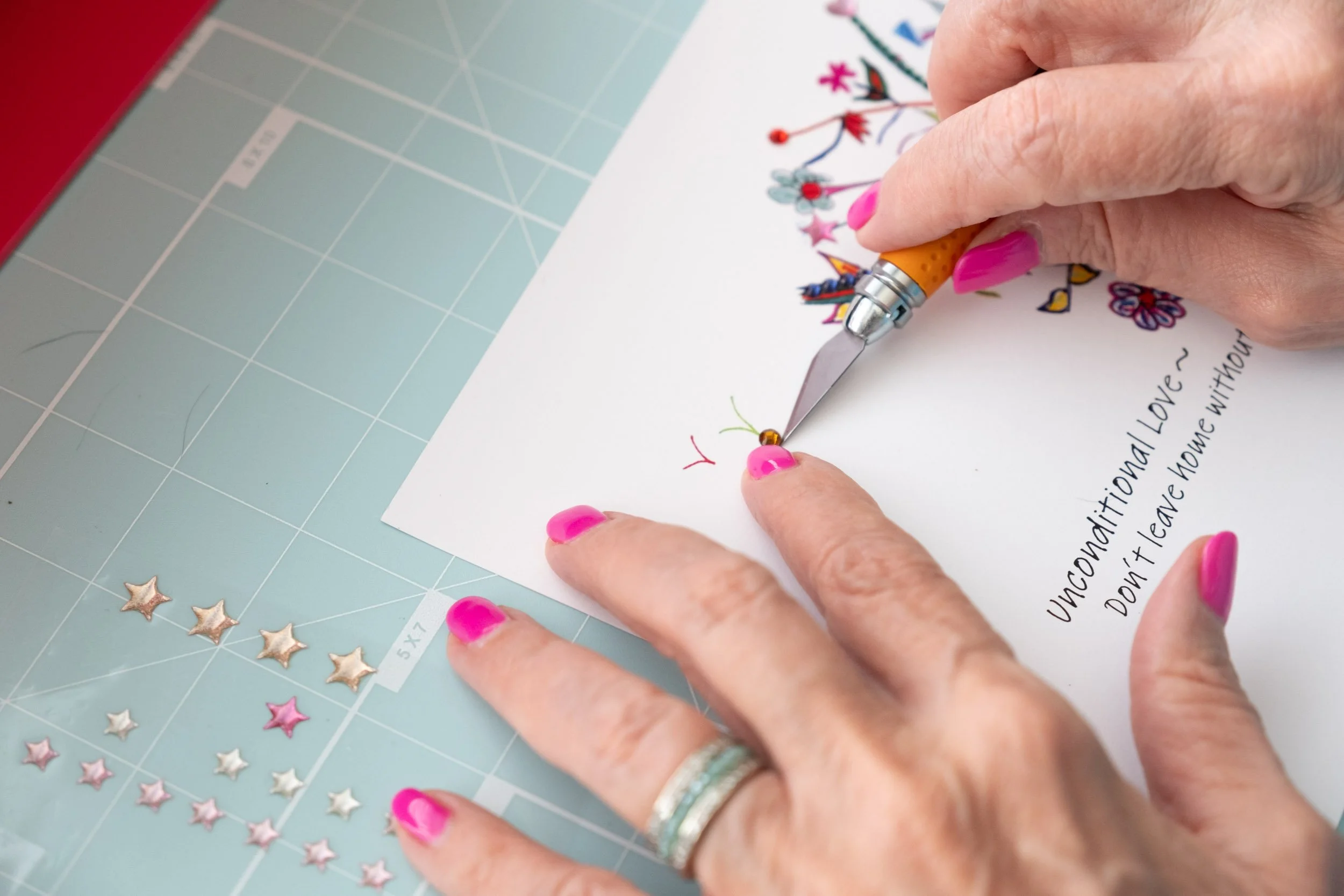 A person with pink painted nails decorating a handwritten card with a small bead and needle, surrounded by star-shaped stickers on a cutting mat.
