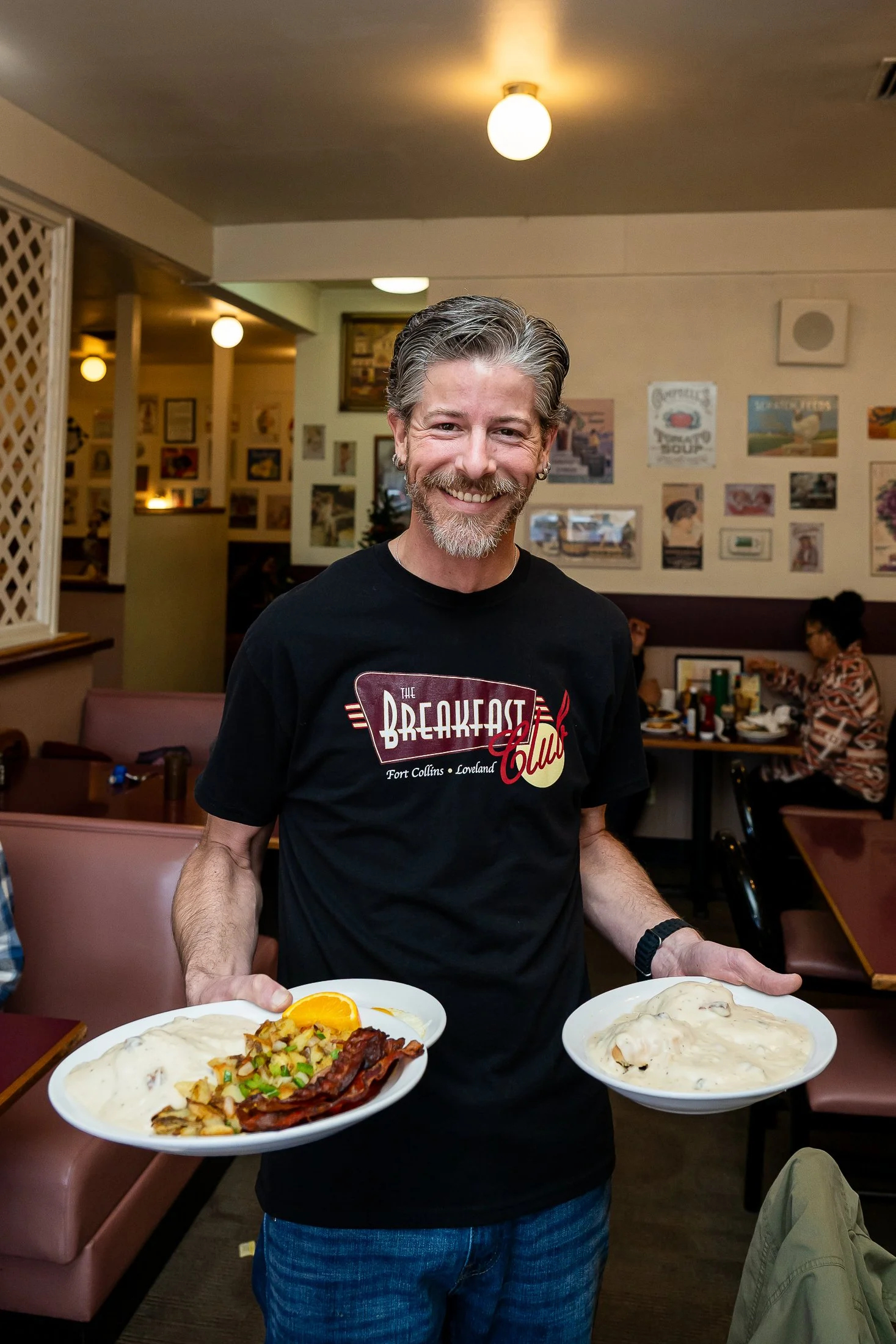 David the general manager of the Loveland Breakfast Club holding two plates of American diner food and smiling at the camera.