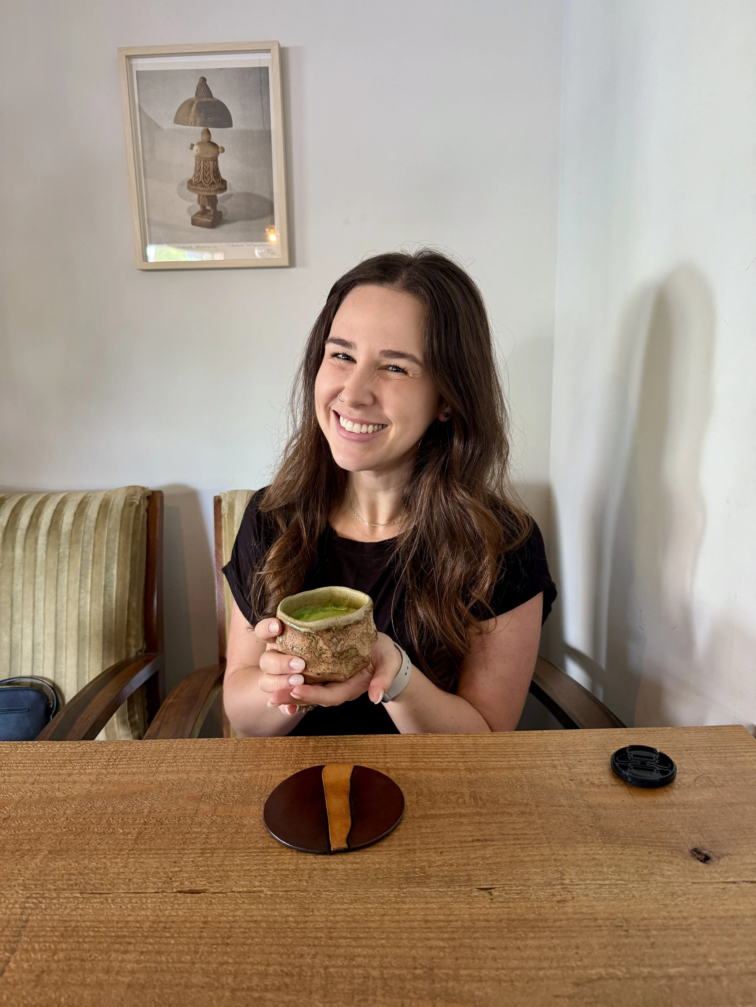 Young woman with long brown hair smiling and holding a green matcha tea in a rustic ceramic cup, sitting at a wooden table in a cafe.