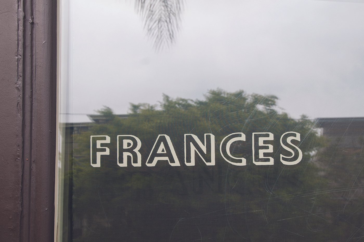 A glass window with the word 'FRANCES' written on it, reflecting trees and an overcast sky outside.