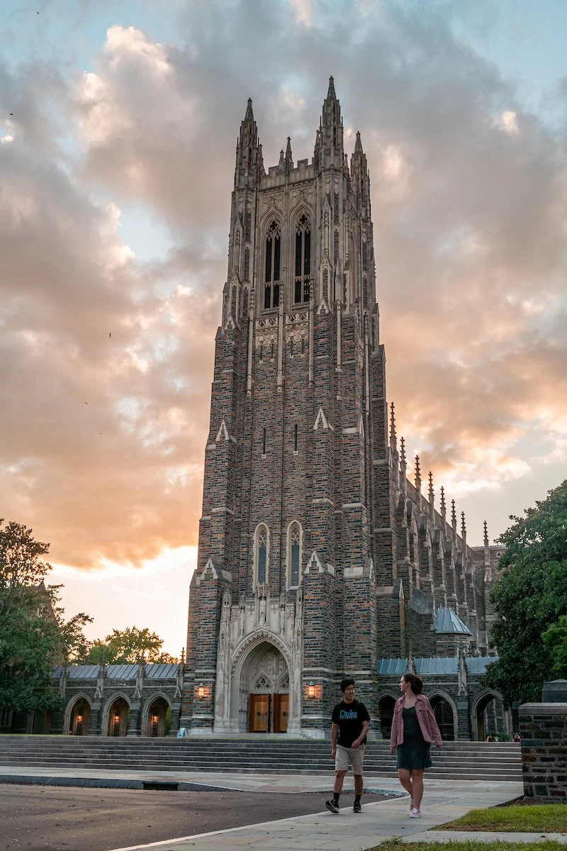 Two students walk by Duke University Chapel under a cloud-dappled sunset