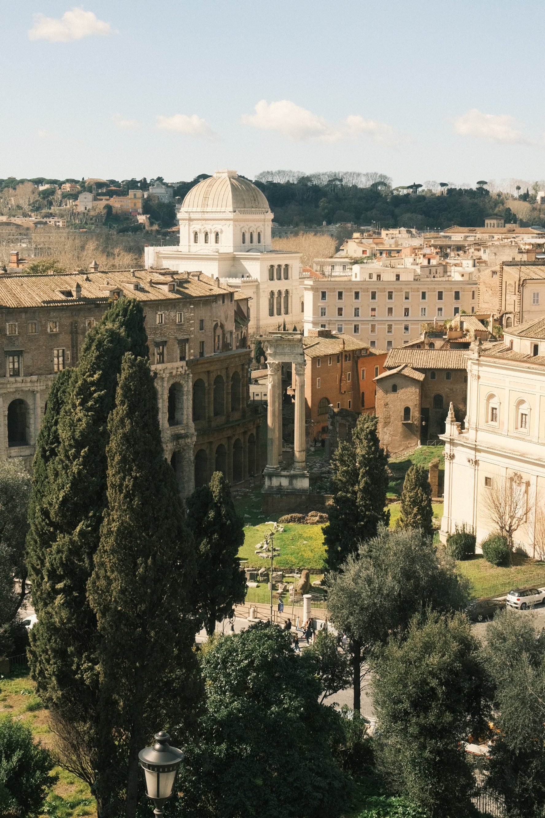 Ancient ruins with columns, surrounded by trees and buildings, under a partly cloudy sky, in a city setting.