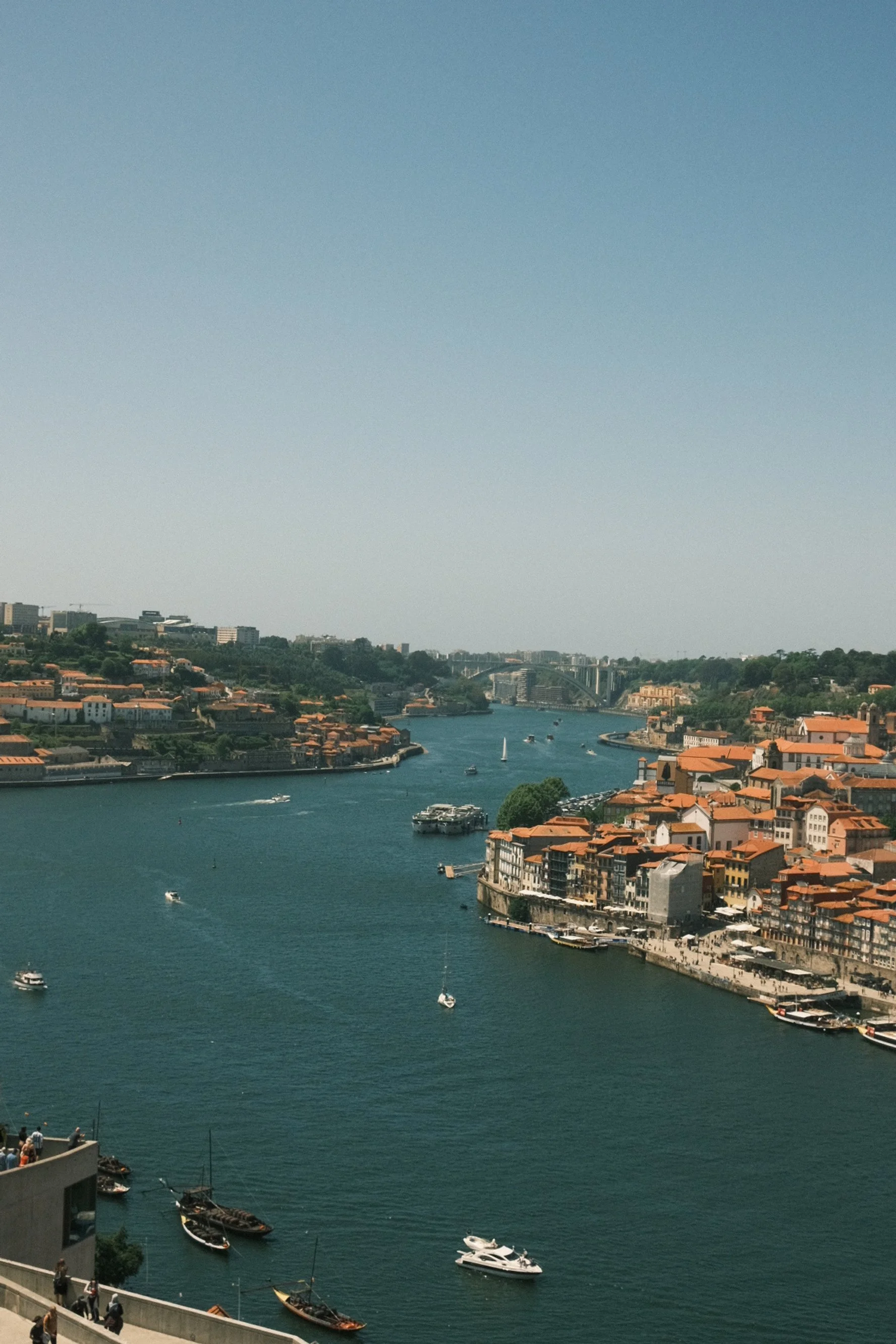 View of a river with boats and sailboats, surrounded by red-tiled roof buildings and green hills in the background.