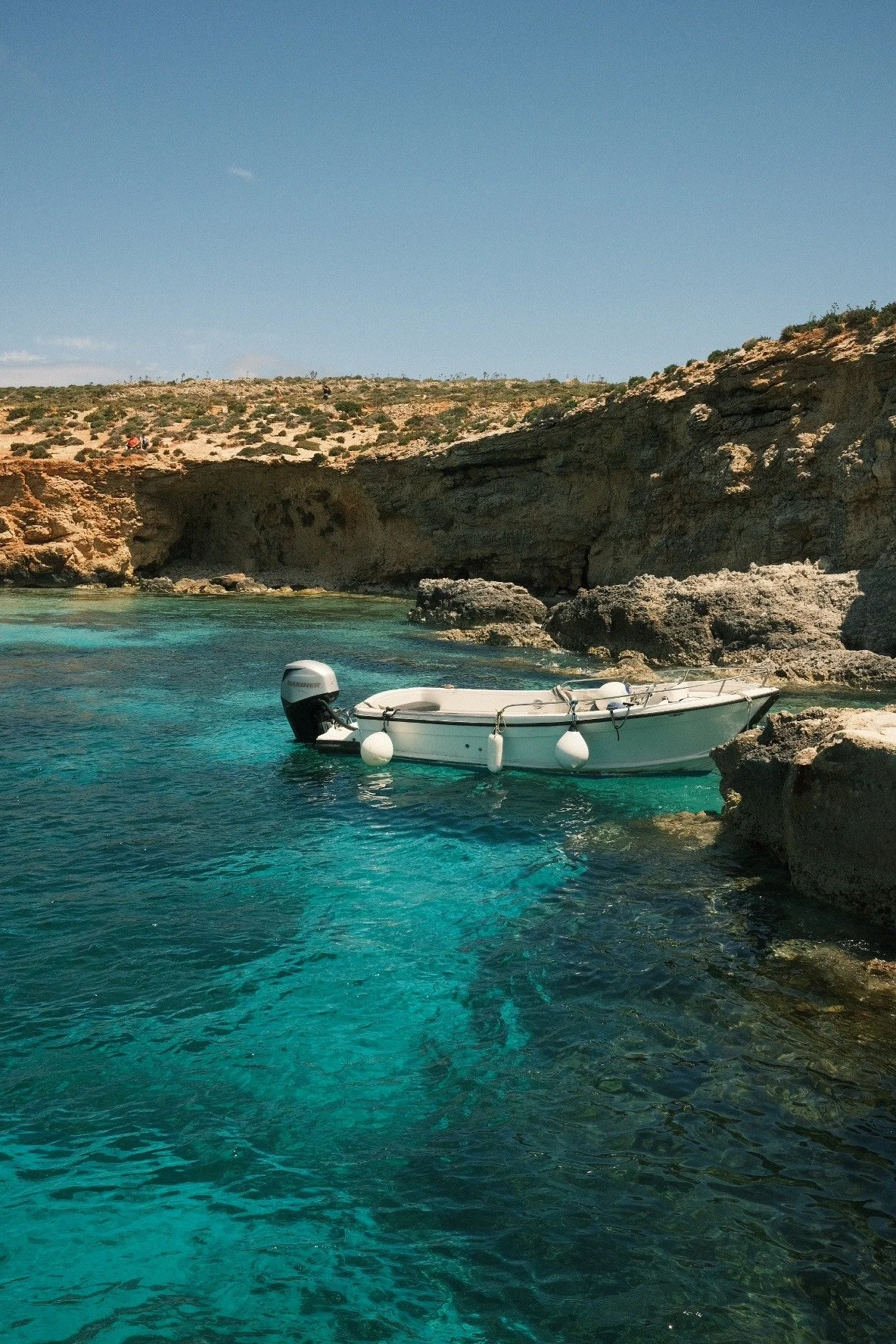 A small white boat is anchored near a rocky shoreline in clear blue water on a sunny day with a dry, sandy hill in the background.