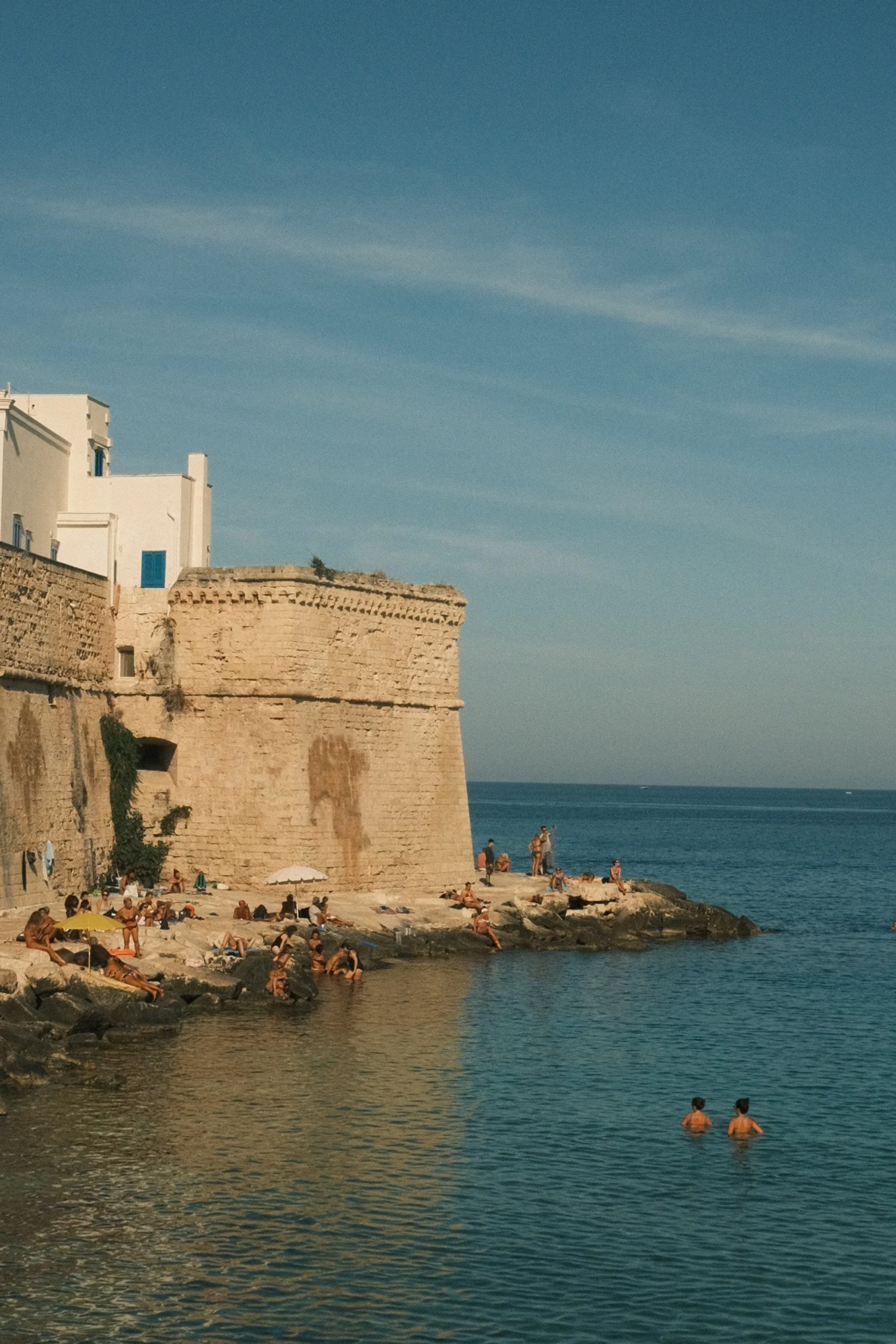 People sunbathing and swimming near a historic stone structure on a rocky coastline under a blue sky.