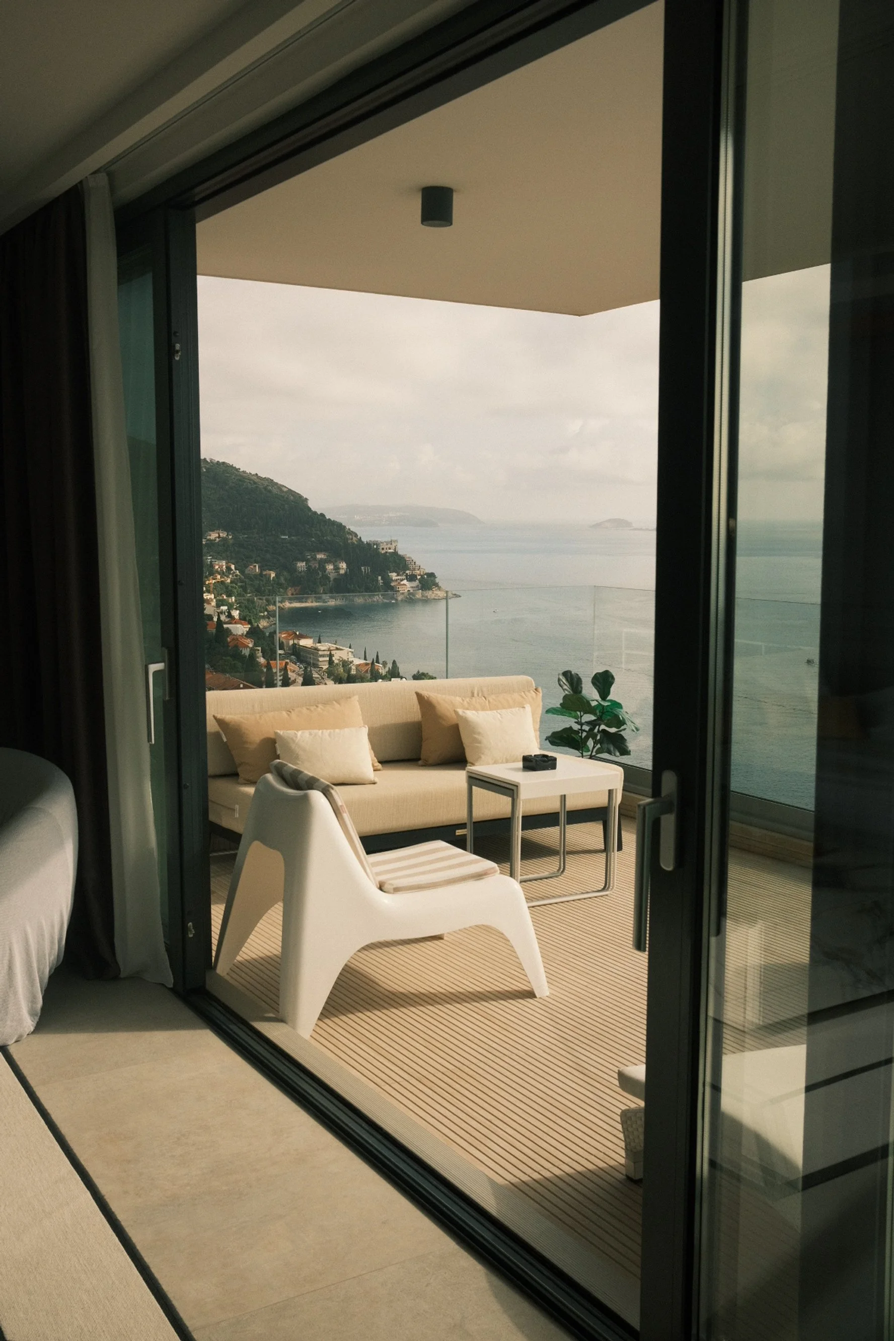 Balcony with beige outdoor sofa, a small table, a white chair, a potted plant with large green leaves, and a view of water and hills in the distance.