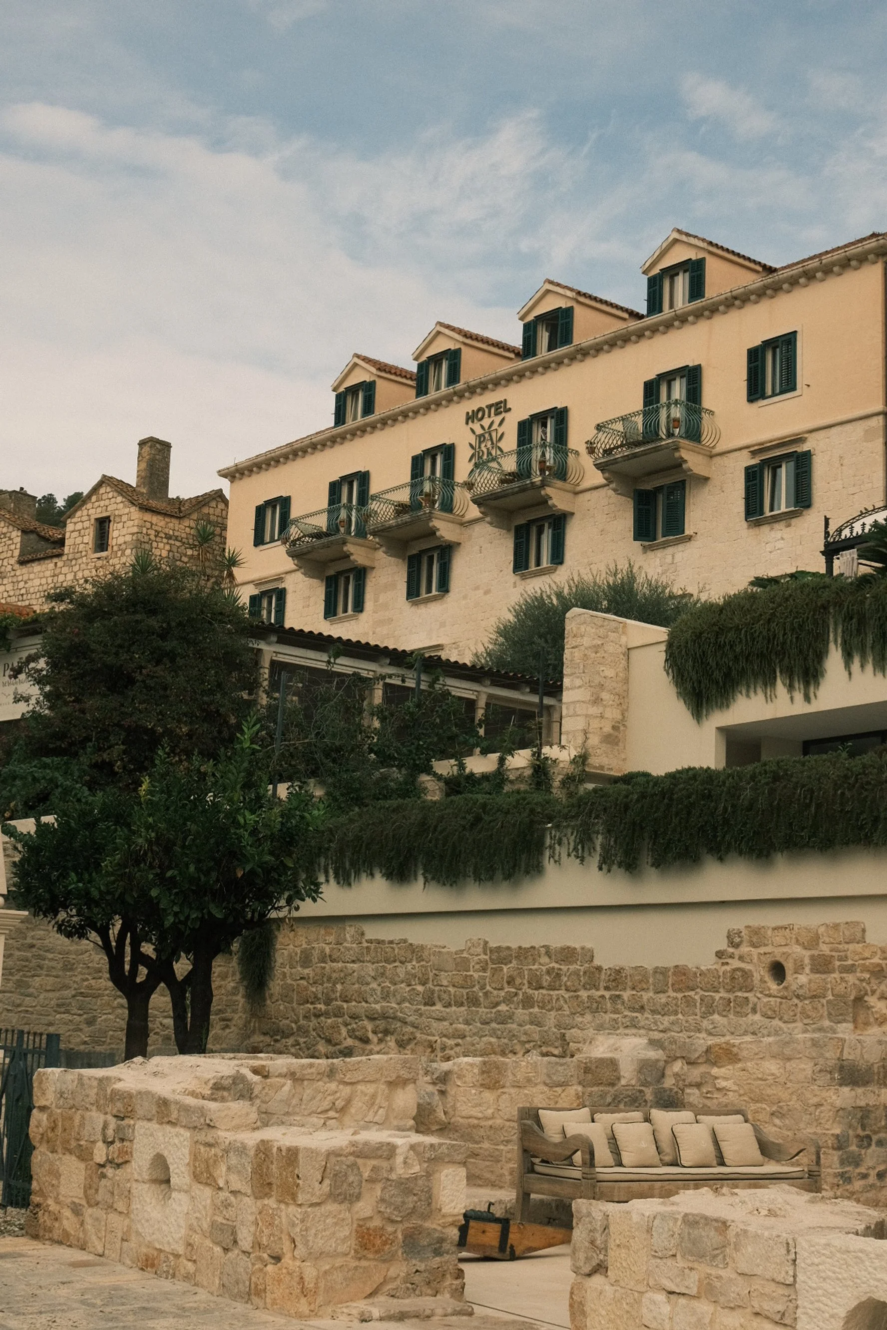 Street view of a multi-story hotel with a sign that reads 'Hotel' and several small balconies, set against a cloudy sky. The foreground features a stone patio with an outdoor sofa and stone walls.