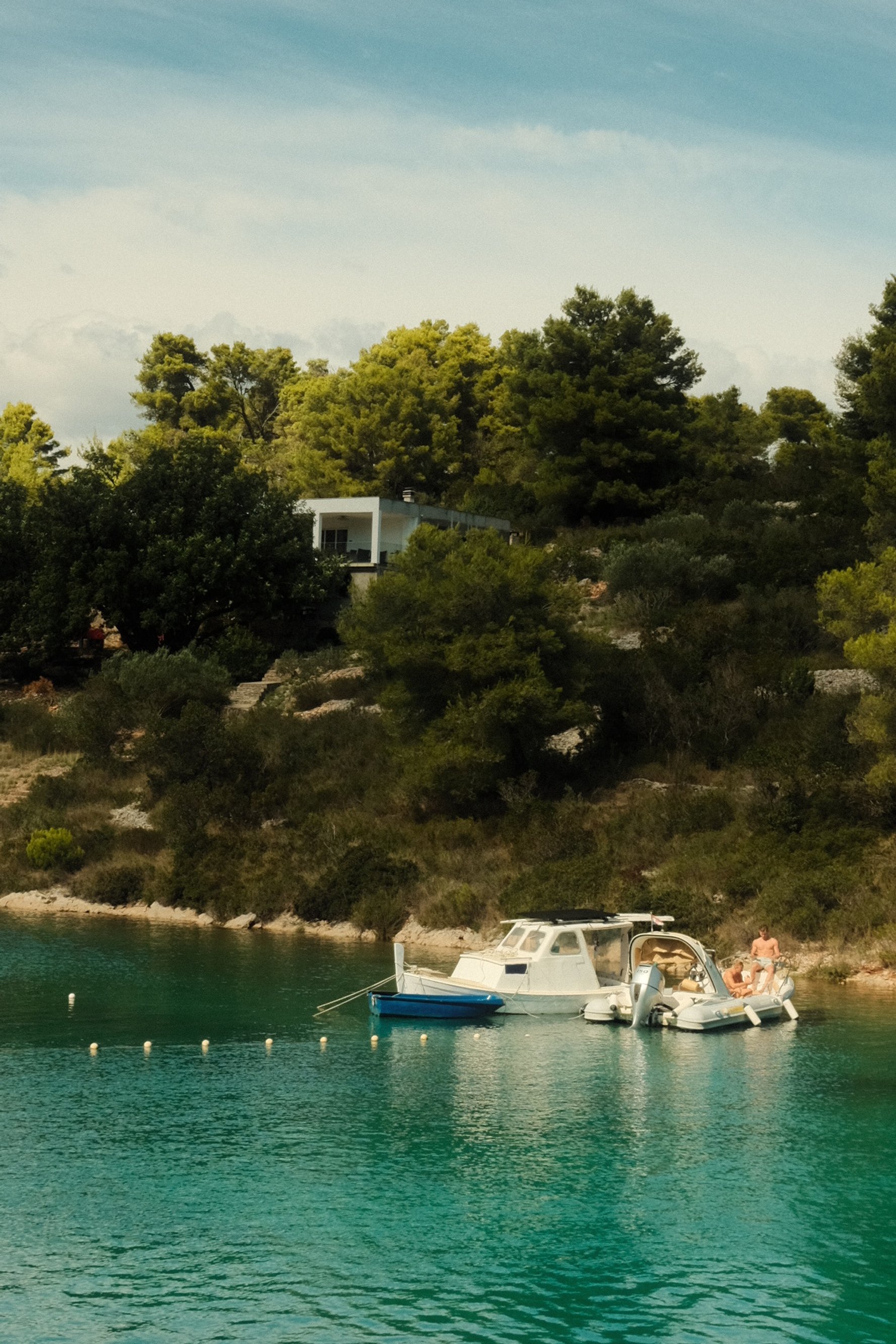 A small motorboat and a pontoon boat docked near a rocky shoreline with deep green trees on a calm body of water, with a modern house on a hillside in the background under a partly cloudy sky.