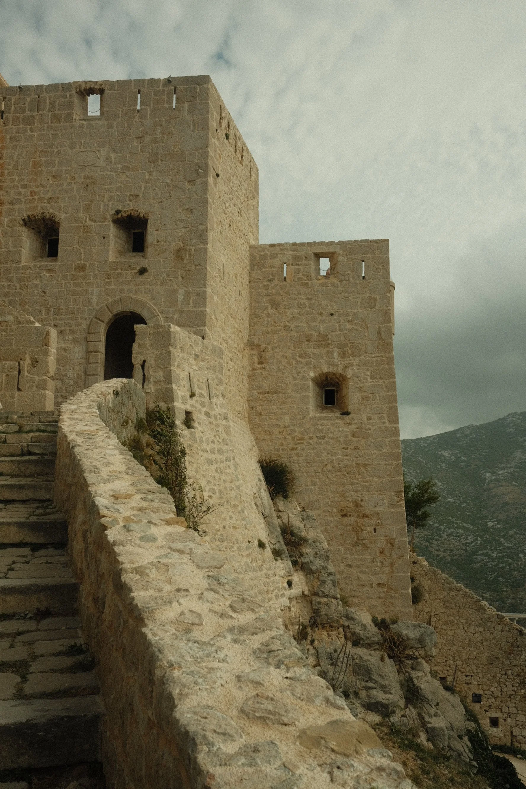 An ancient stone castle with stairs leading to an arched entrance, situated on a rocky hillside under a cloudy sky.