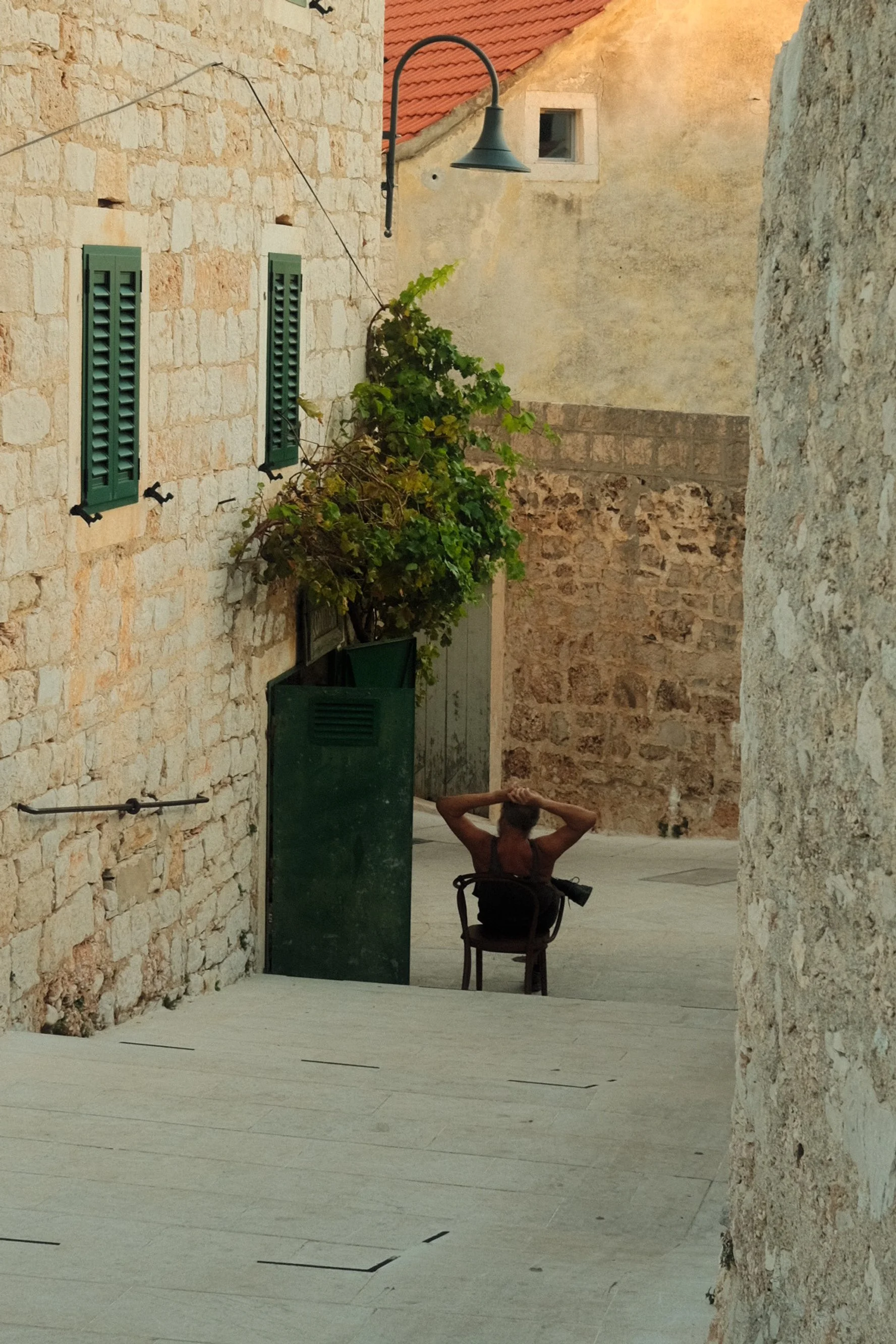 Person sitting on a chair with arms behind head in a narrow stone alleyway with old buildings, green shutters, and a streetlamp