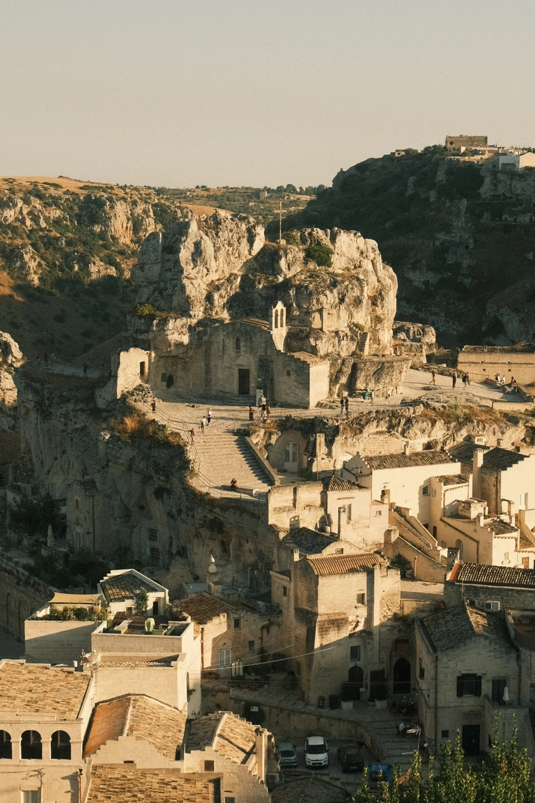 Historic cave dwellings and buildings in a rocky hillside town, with stairs and people walking, under a clear sky at sunset.