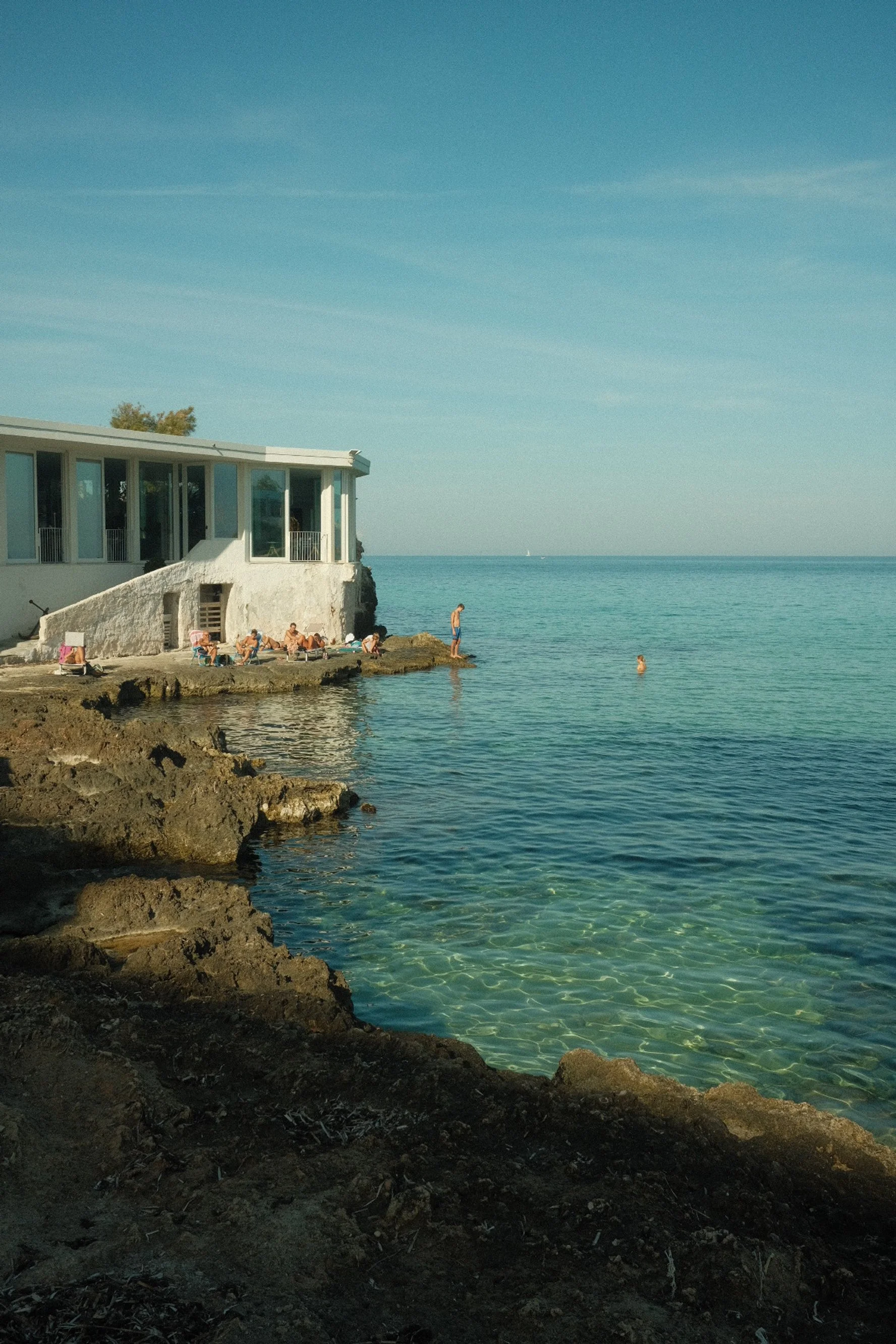 People relaxing and swimming near a white building on the rocky shoreline of a clear blue ocean on a sunny day.