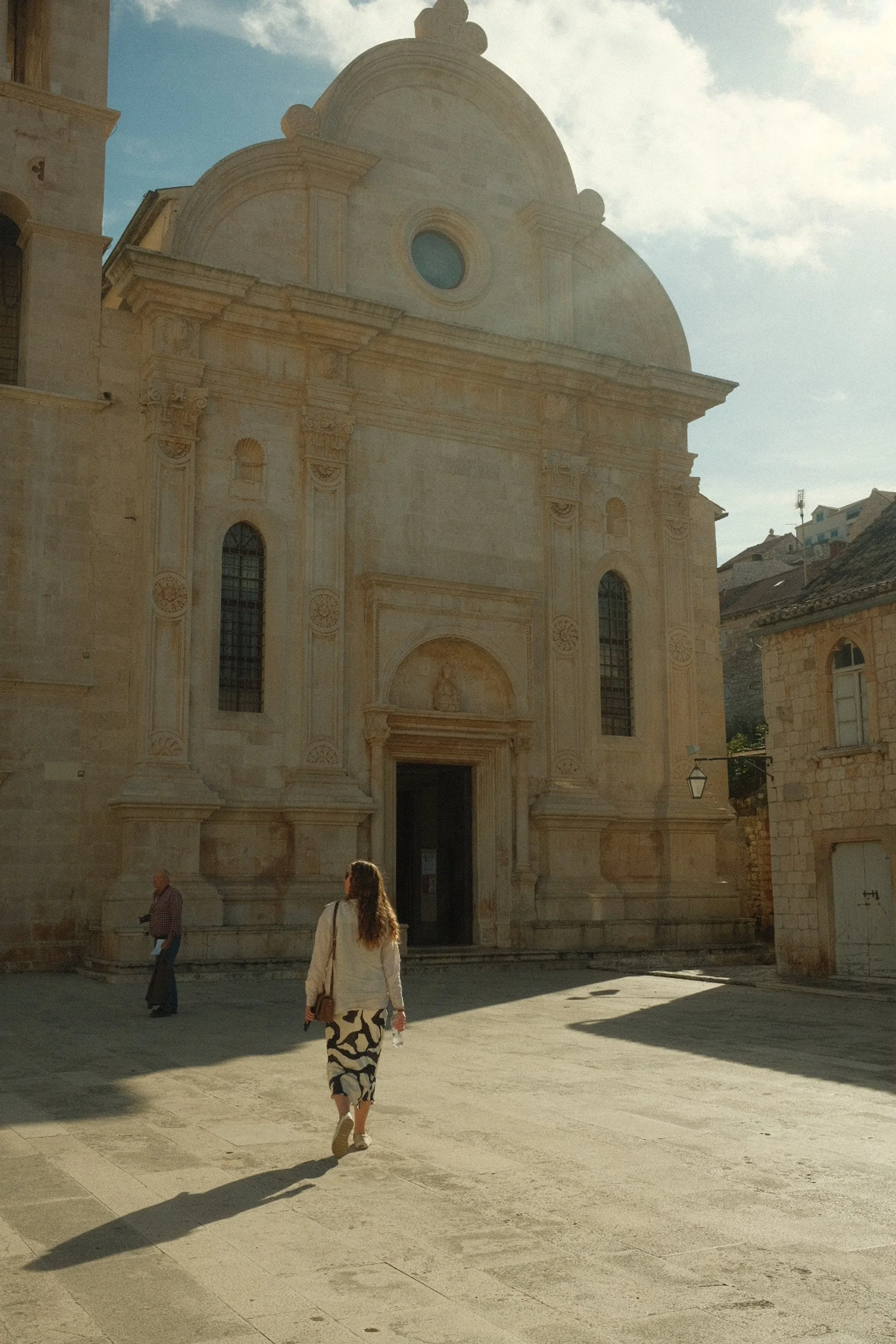 A woman walking towards a large stone church with baroque architectural details, with sunlight creating long shadows on the ground.