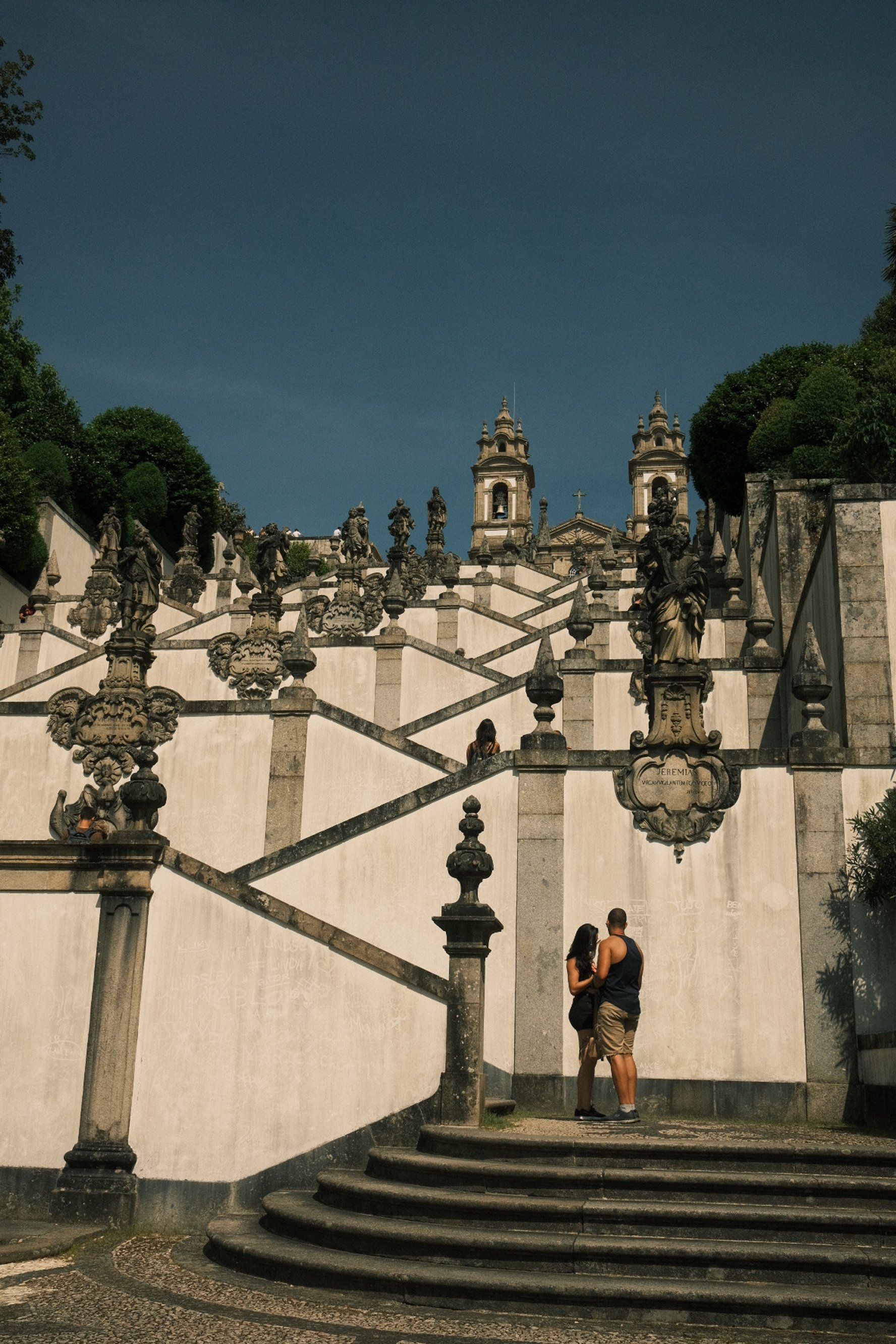 People standing on steps at the bottom of an ornate, historic staircase with statues and towers at the top, against a dark blue sky.