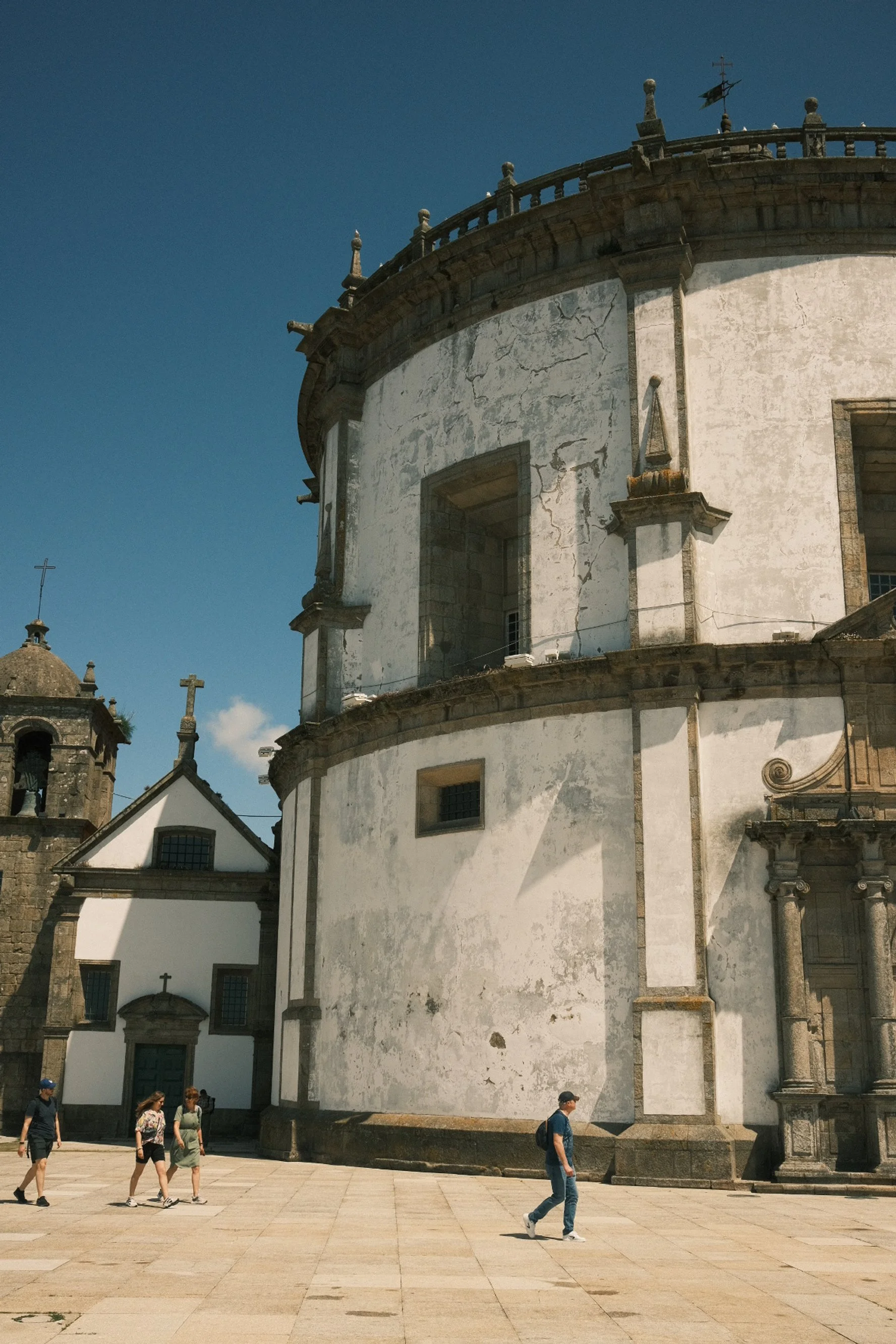 Historic church with a rounded, weathered white facade and small group of tourists walking nearby, under a clear blue sky.