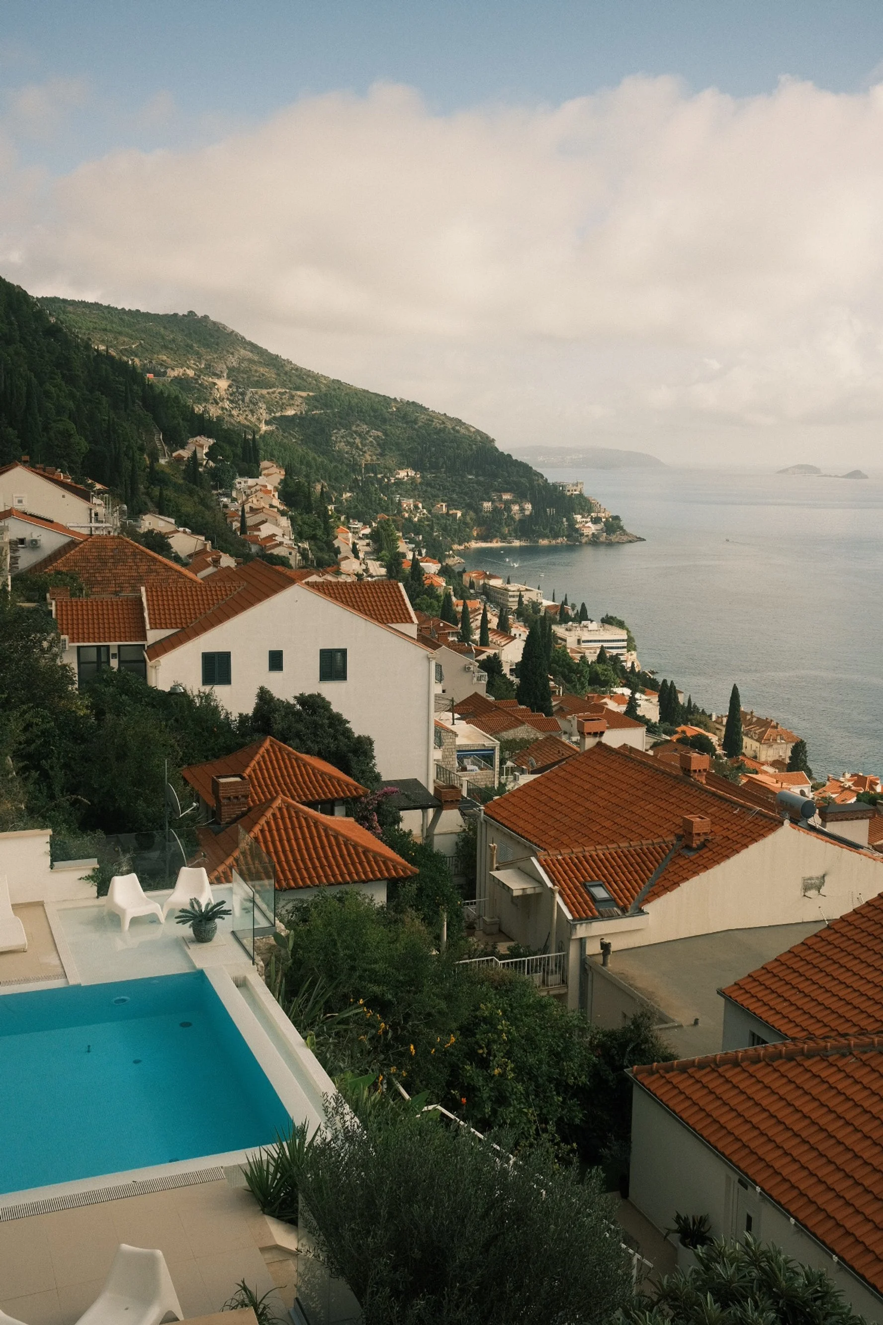 View of houses with red tile roofs on a hillside overlooking the sea, with a swimming pool in the foreground and a partly cloudy sky.