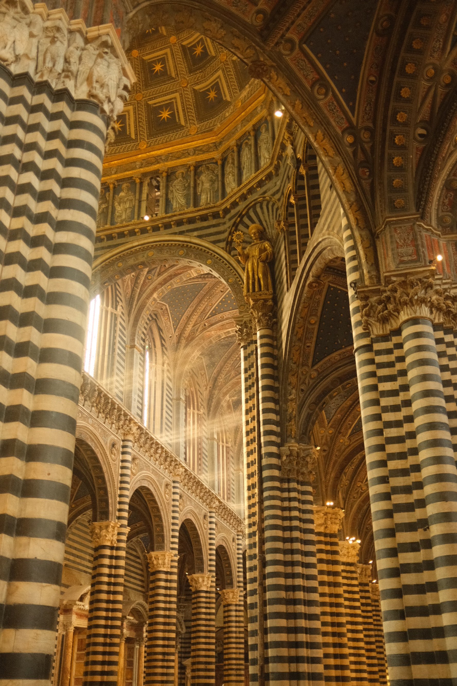 Interior of a cathedral with black and white striped columns, ornate arches, gold statues, and a decorated ceiling with blue and gold accents.