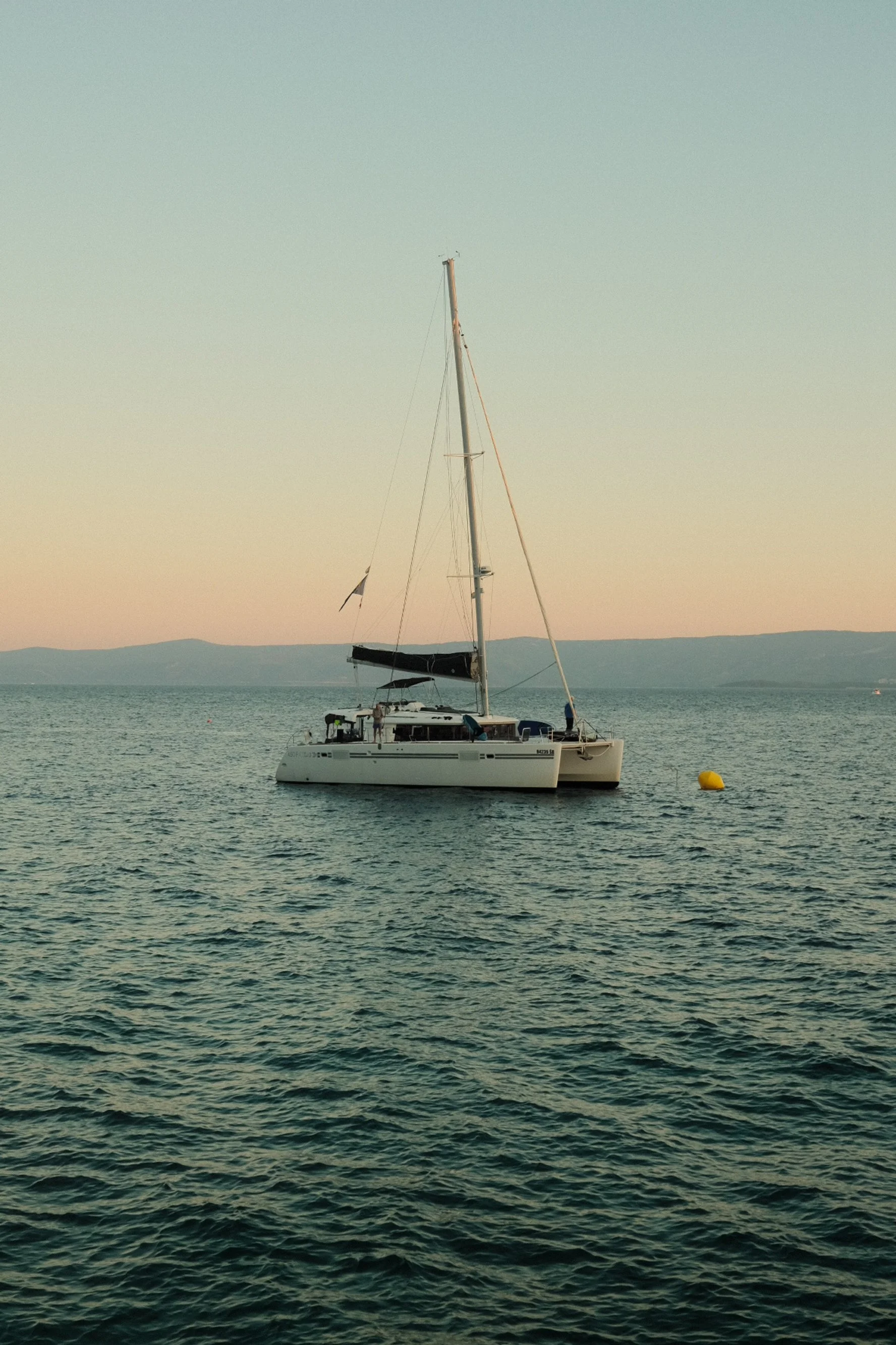 A sailboat floating on calm water near a yellow buoy, with a distant shoreline and hills under a clear sky at sunset.