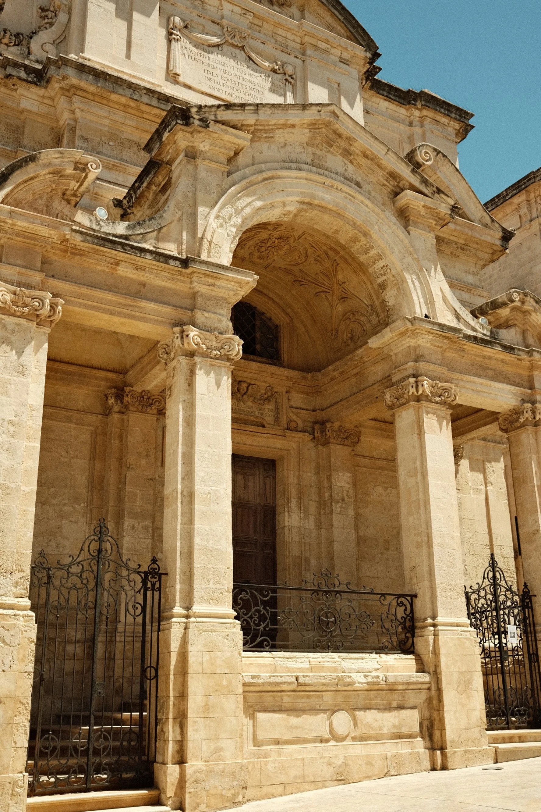 The front facade of a historic stone church with columns, a wooden door, wrought iron gates, and decorative architectural details under a clear blue sky.