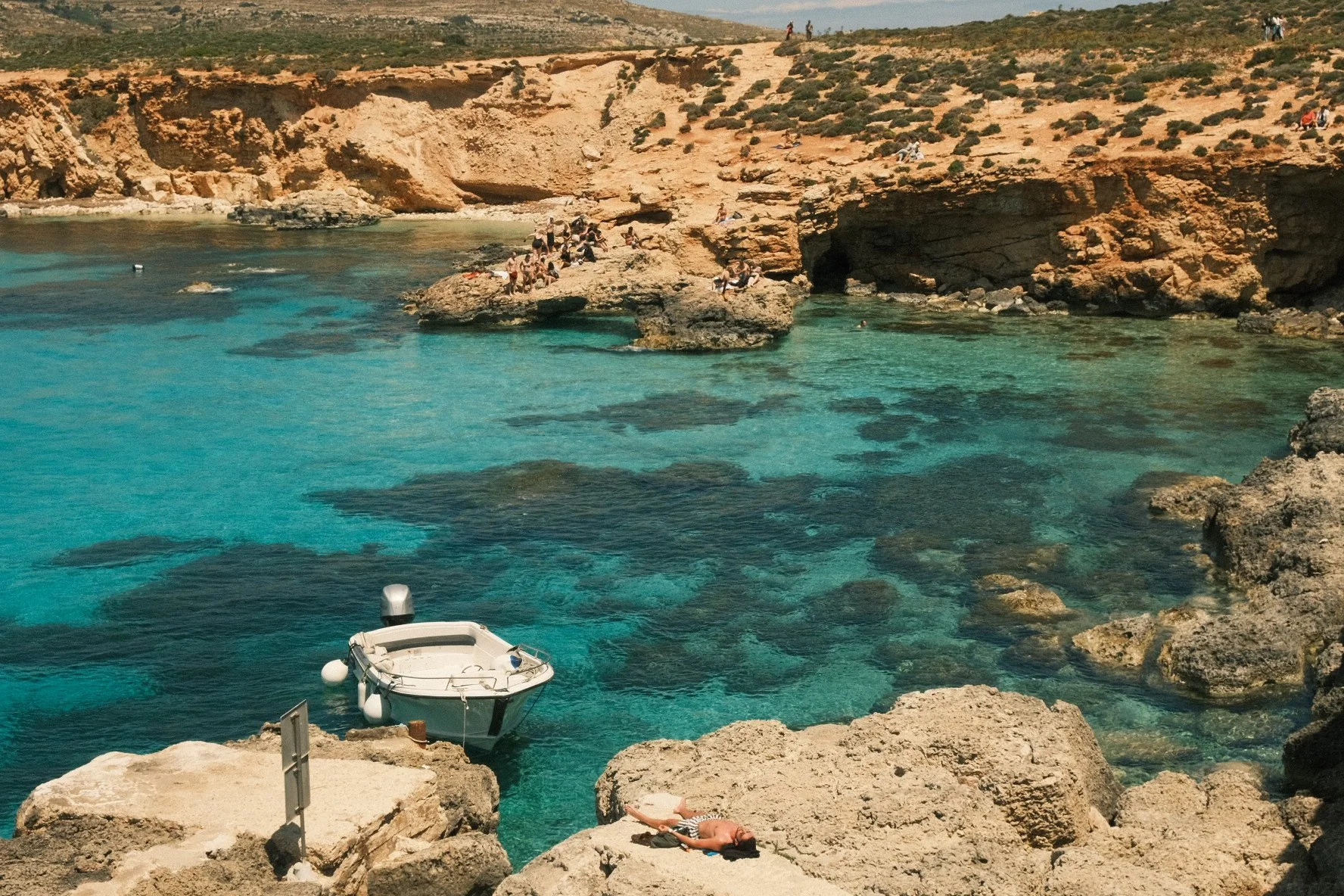 A scenic coastal area with turquoise water, rocky cliffs, and people relaxing and sunbathing on rocks and sitting by the water.