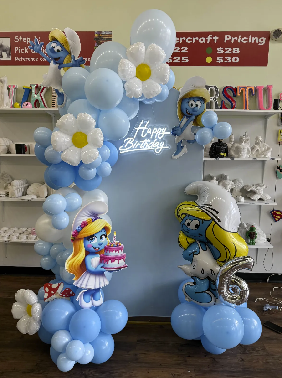 Balloon decoration featuring Smurfs, including Smurfette and other characters, with a "Happy Birthday" sign, blue and white balloons, and decorative flowers, for a birthday celebration.