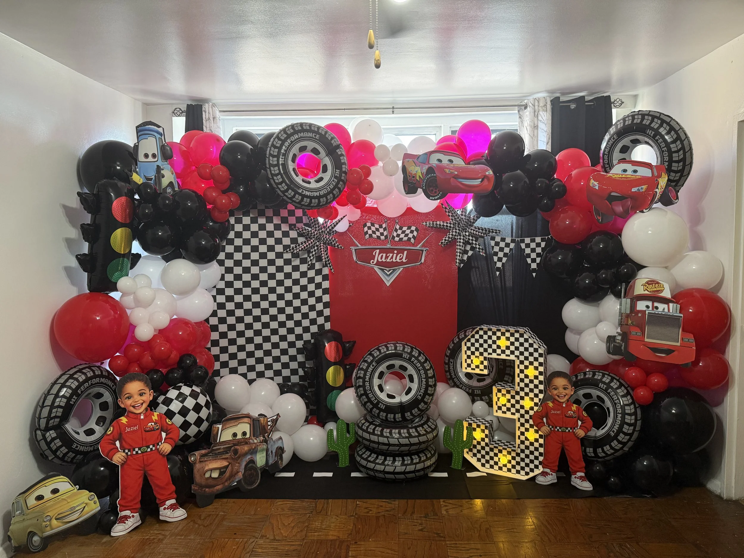 Colorful decorations for a Lightning McQueen-themed birthday party with balloons, cardboard cutouts of children, cars, tires, checkered flags, and a large number 3, with a red banner that says "Jaziel" at the center.