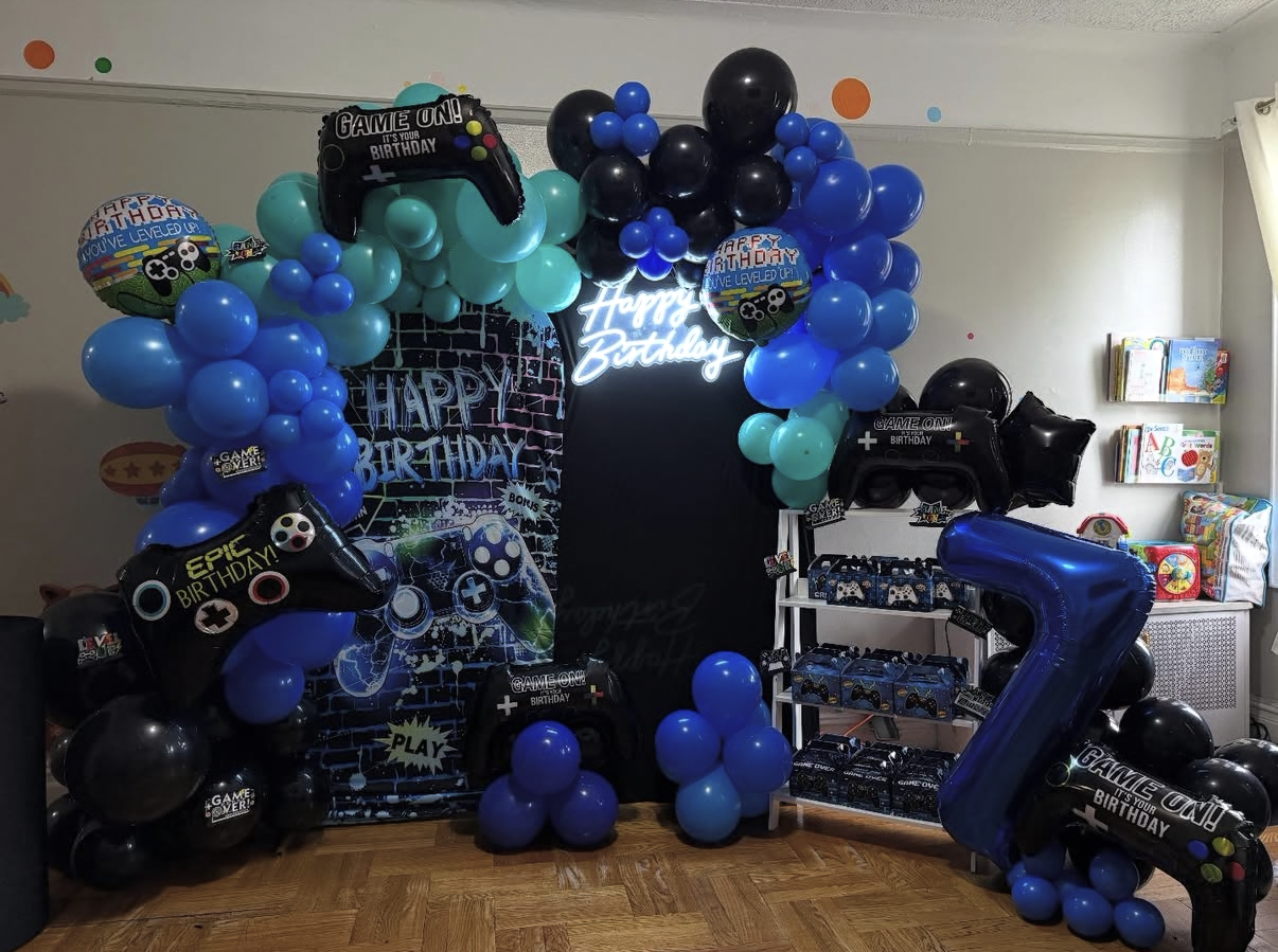 Game-themed birthday party decorations with black, blue, and teal balloons, a large gaming controller balloon, a large number one balloon, a chalkboard backdrop with birthday message, and shelves with party favors.