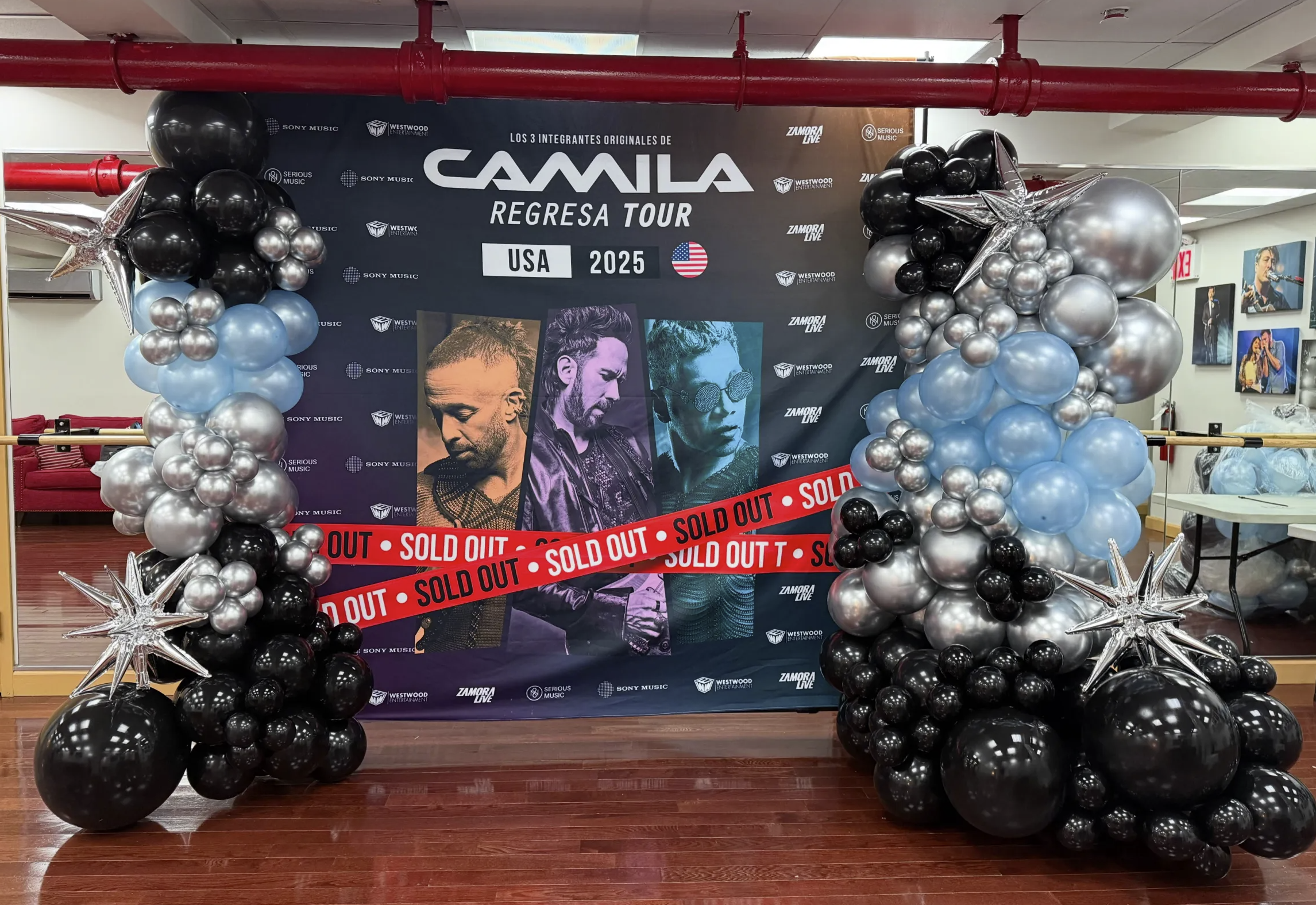 A decorated backstage area with a large poster of the musical group Camila, featuring three members, and balloons in black, silver, and light blue. The balloons are arranged on either side of the poster, with a large silver star balloon on each side, and a red 'sold out' tape across the front.