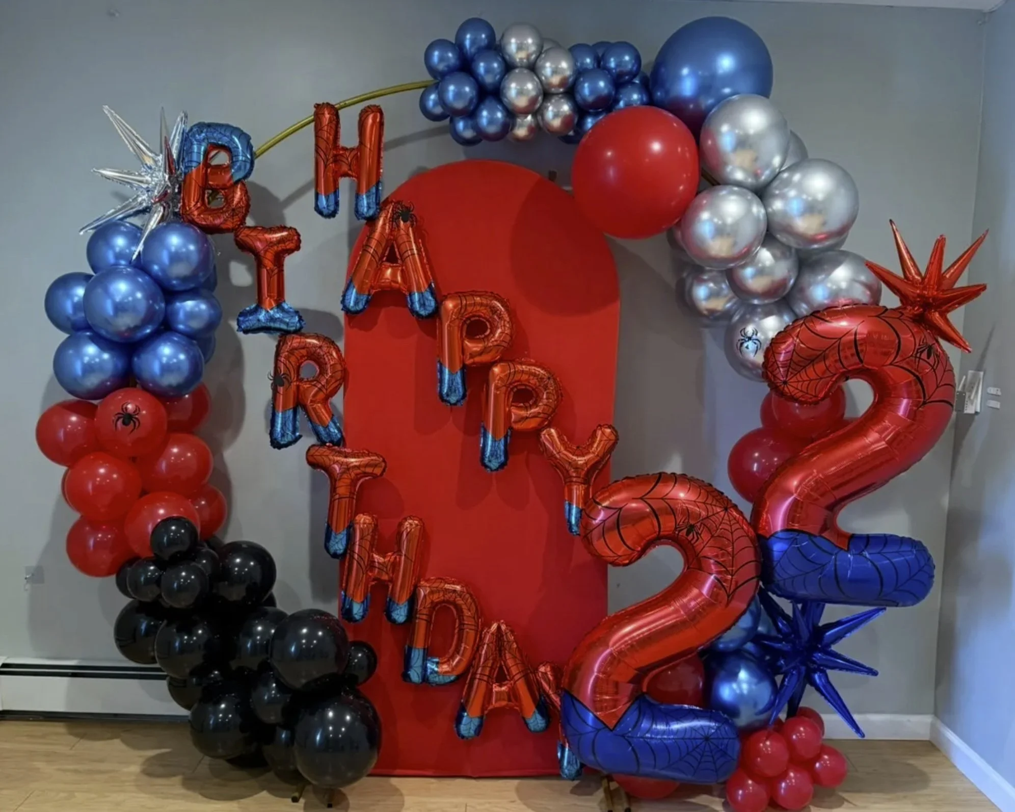 A Halloween-themed balloon decoration with red, blue, black, and metallic silver balloons, spelling out 'Happy Birthday' in stylized letter balloons. Large numbers '2023' with web design on the right side. Red backdrop in the center.