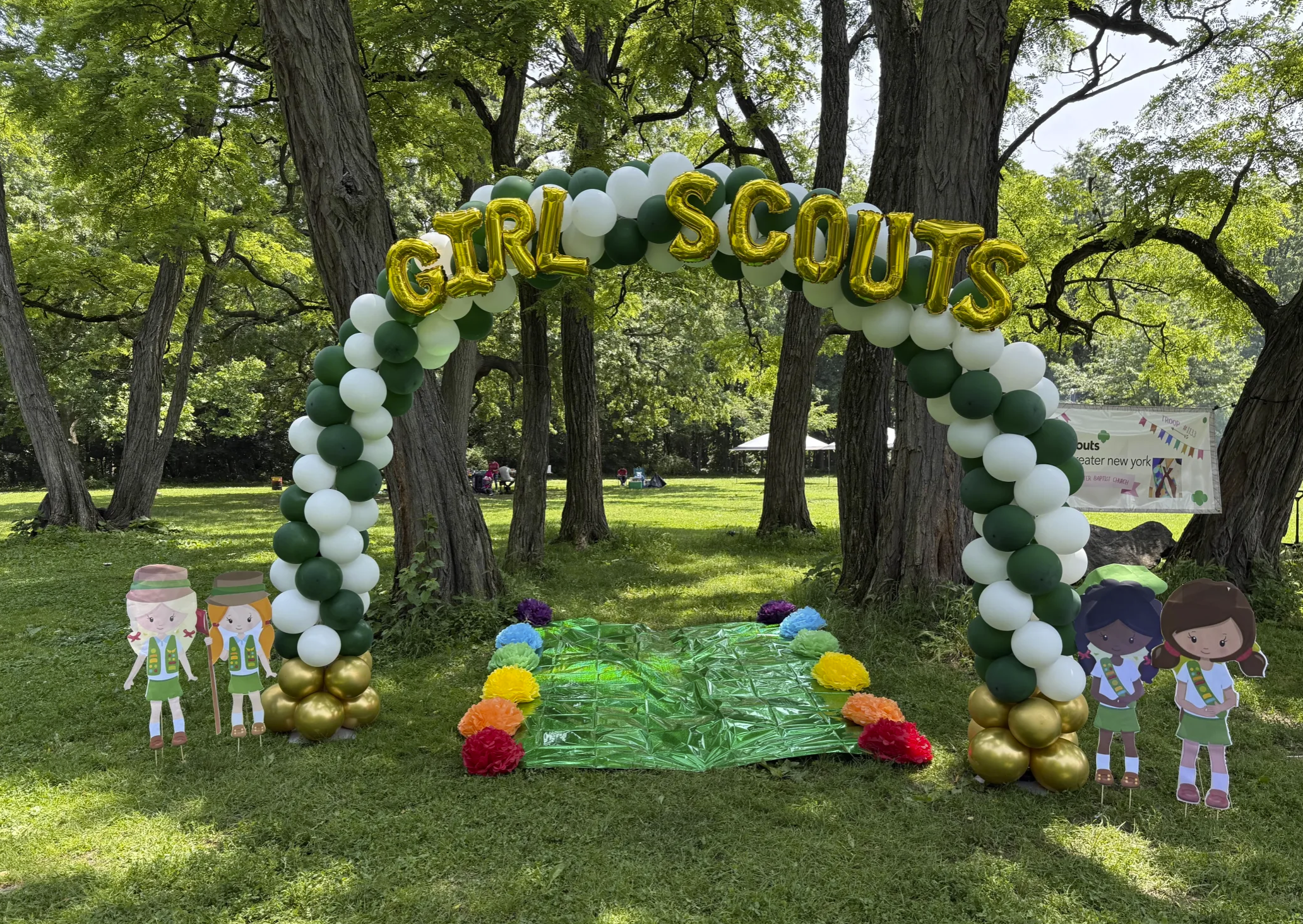 Decorative arch made of green, white, and gold balloons with gold letter balloons spelling 'GIRL SCOUTS' at the top. There are cartoon cutouts of girl scouts on either side of the arch. The ground underneath is covered with rainbow-colored tissue paper pom-poms and a shiny green mat. The setting is outdoors in a park with trees and grass.