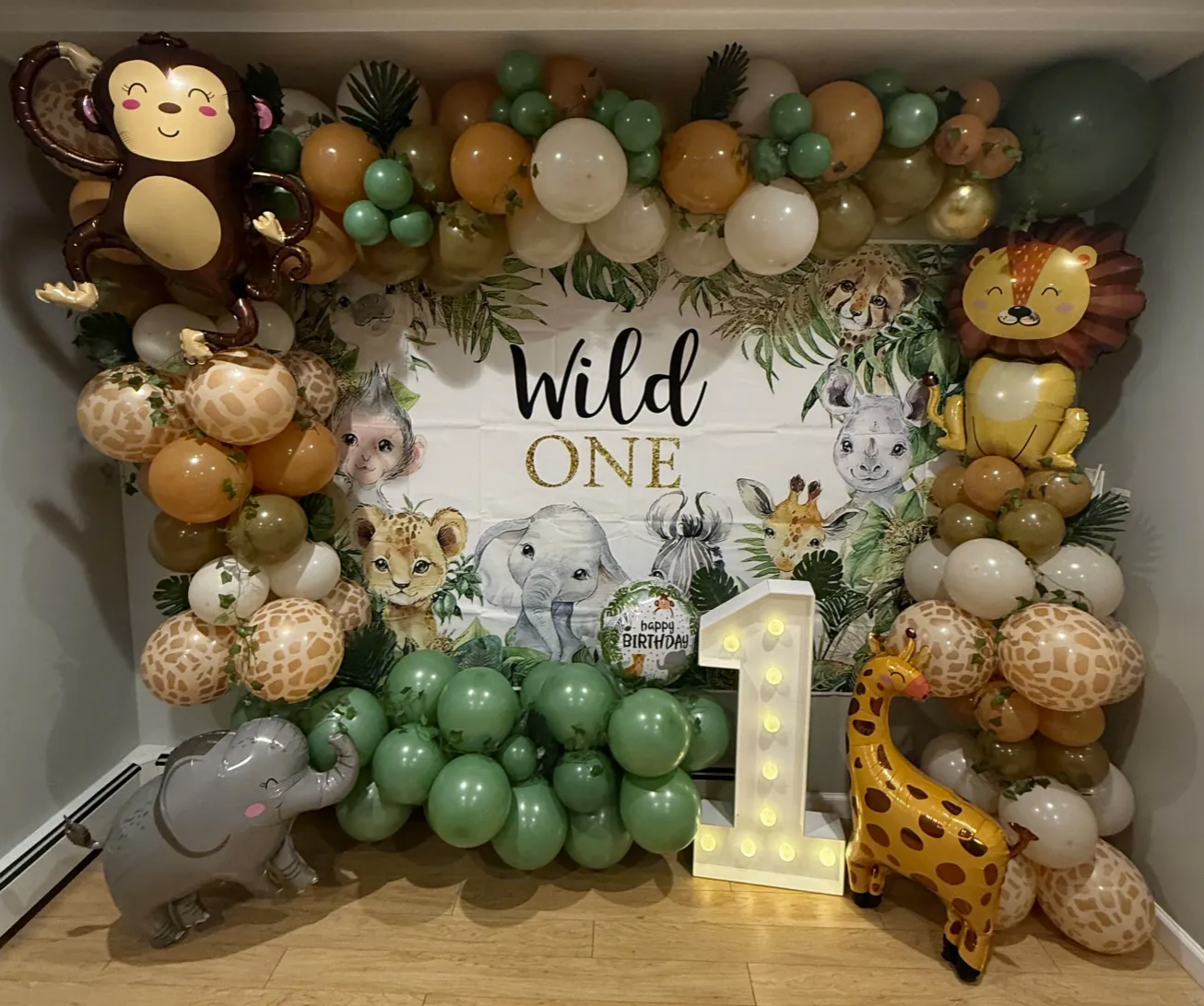 Decorative jungle-themed birthday party setup with balloons featuring animal prints and cartoon animal balloons, a large marquee number '1', and a backdrop that reads 'Wild One' with illustrations of jungle animals, leaves, and a birthday message.