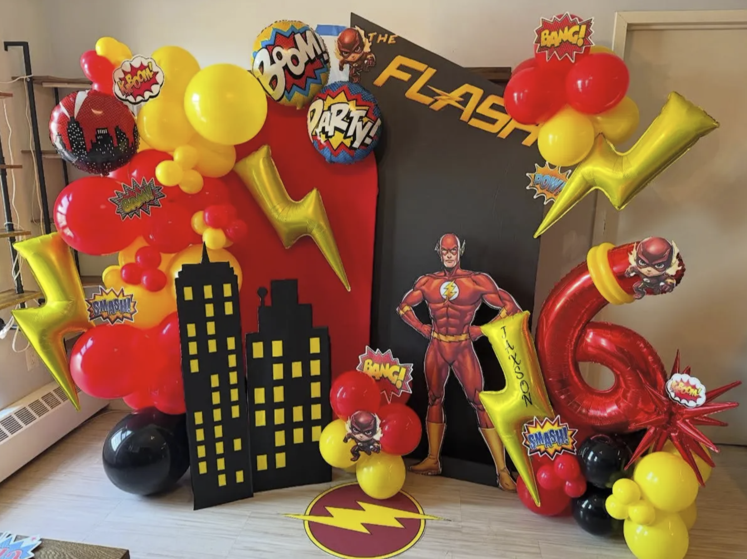 Superhero-themed birthday decoration with balloons, cityscape cutout, and superhero cutouts, featuring The Flash character.