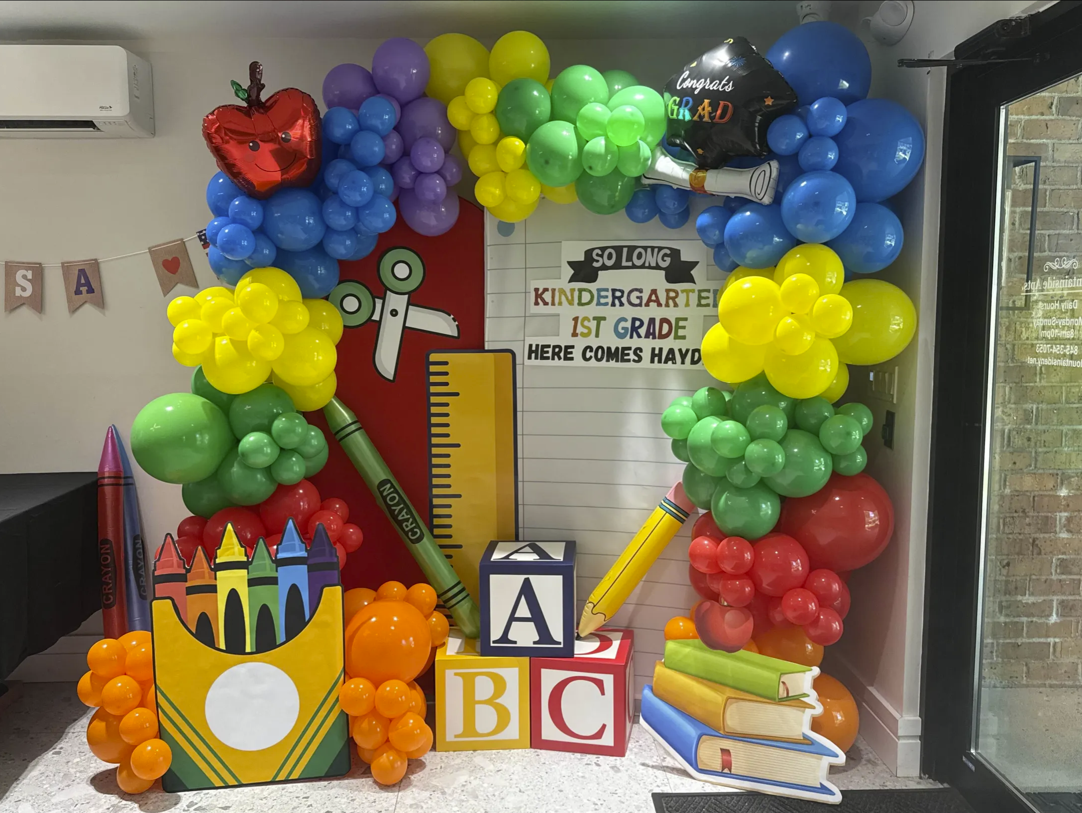 Colorful balloon decoration with a school theme, including balloons shaped like a crayon box, alphabet blocks, books, and pencils. There is a sign welcoming kindergarten and first-grade students, and a sign with a ruler and scissors images.