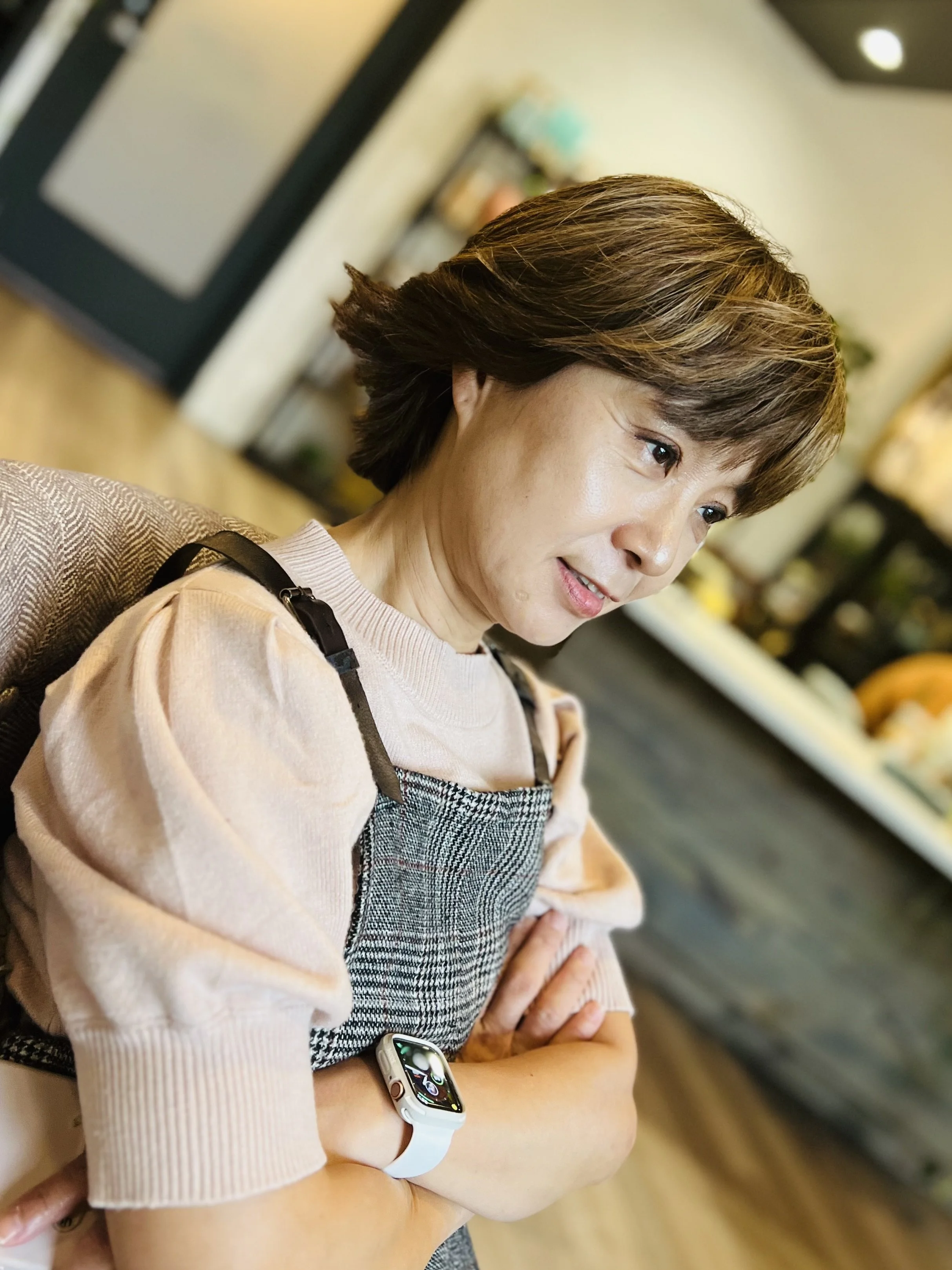 A woman with short hair posing in front of a brick wall, wearing a dark top and a light-colored apron.