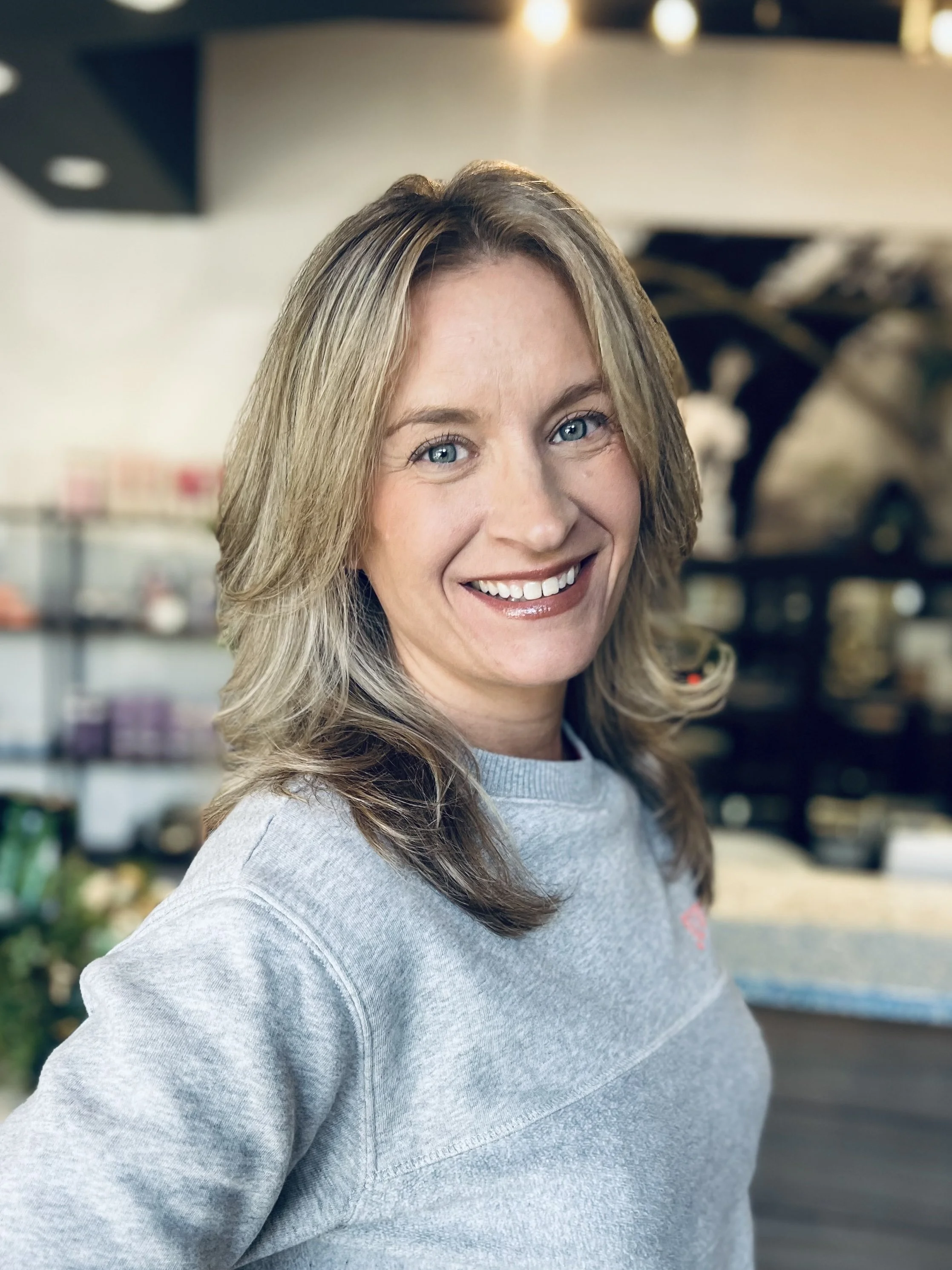 A woman with blonde wavy hair smiling, wearing a white sweater with ruffles, standing outside near a house with white siding and a window.