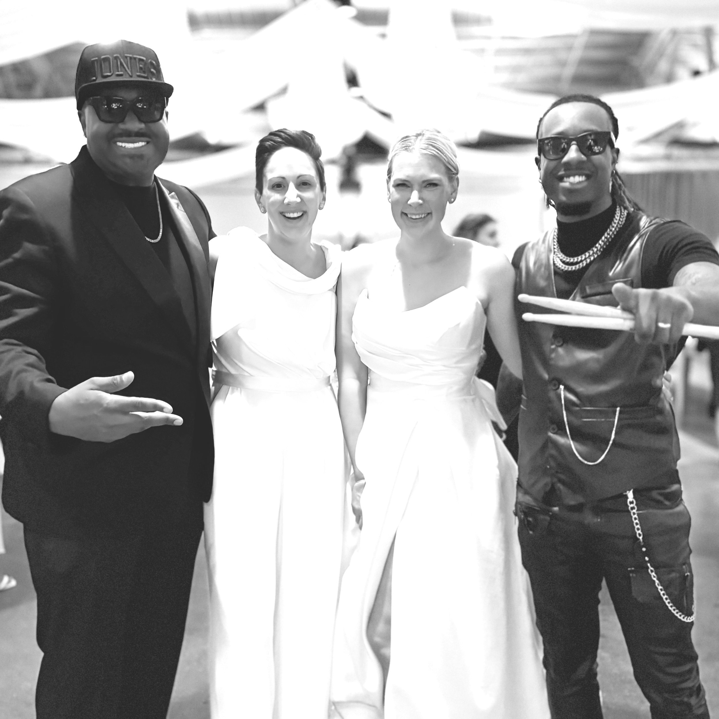 The Rich Rockstars Hybrid Band at a social event, two women in white dresses and two men in dark clothing and sunglasses, standing outdoors with umbrellas in the background.