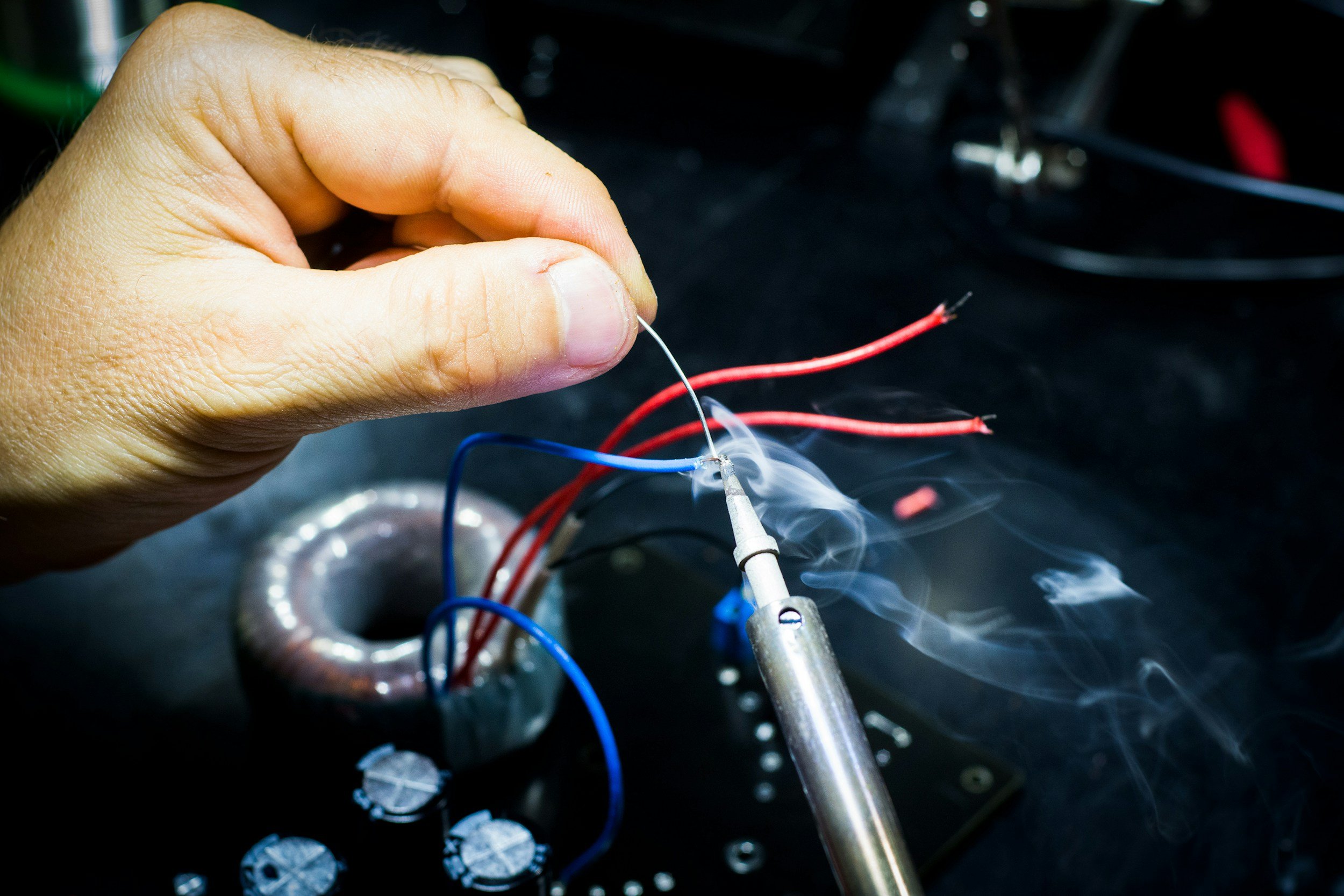 A person soldering red and blue wires with a soldering iron, creating a connection with smoke rising from the heated metal.