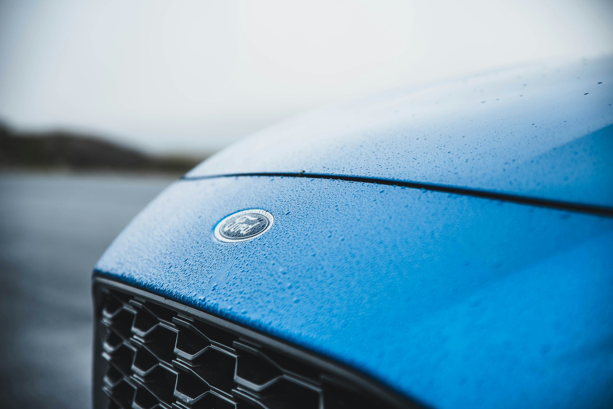 Close-up of a blue Ford car hood with rain droplets and the Ford emblem.