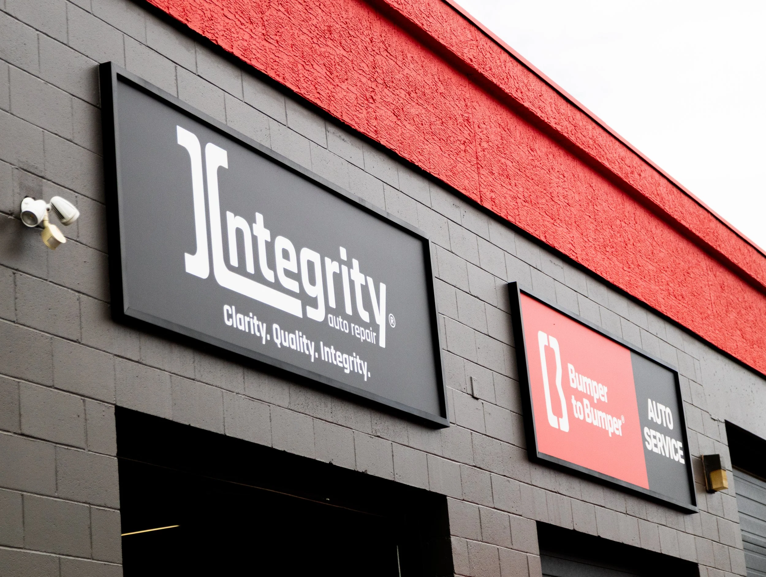 Two signs on the exterior wall of an auto repair shop. The left sign says 'Integrity auto repair' with the slogan 'Clarity. Quality. Integrity.' The right sign says 'Bumper to Bumper auto service' with a red and gray background and a white icon of a