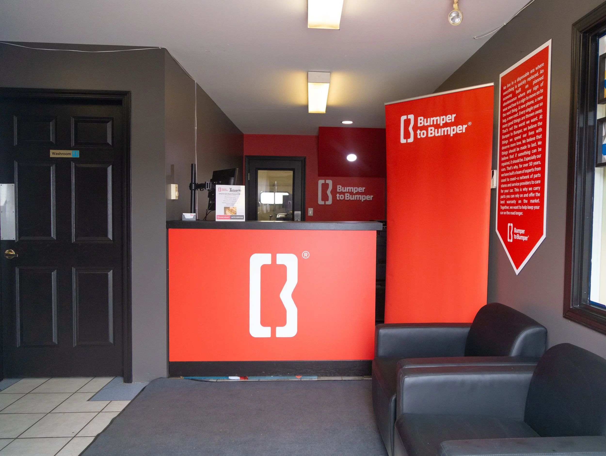 Interior of a car service shop with a red front desk featuring the Bumper to Bumper logo, a black door labeled Washroom, black leather armchairs, and a wall-mounted red banner with the company logo and information.